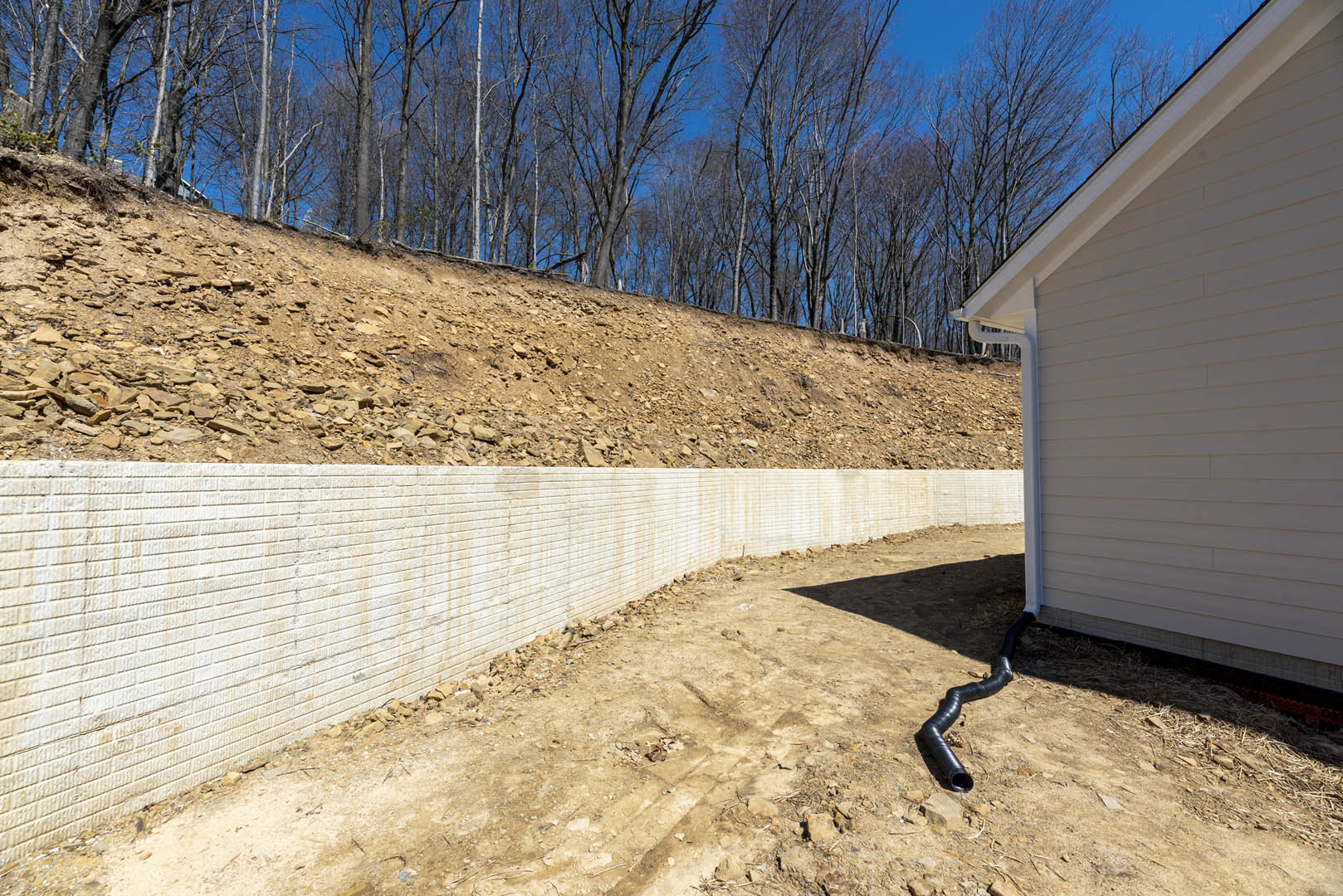 White brick exterior wall of a house with a black pipe running along the ground, bordered by rocks and a dirt hill, with a group of trees on the hillside in the background.