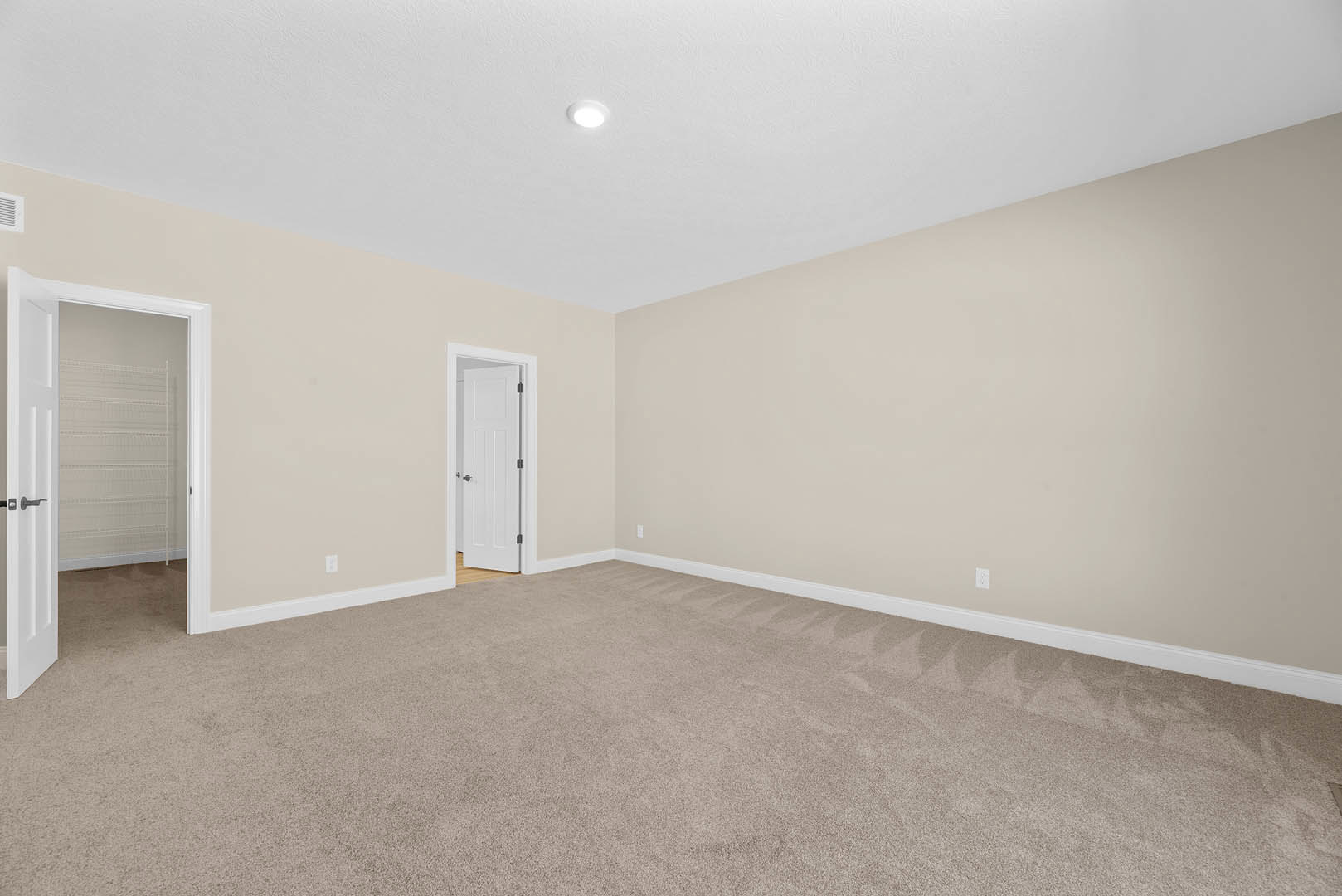 Beige carpeted room with white door, white walls, ceiling light fixture, and wall-mounted white shelf