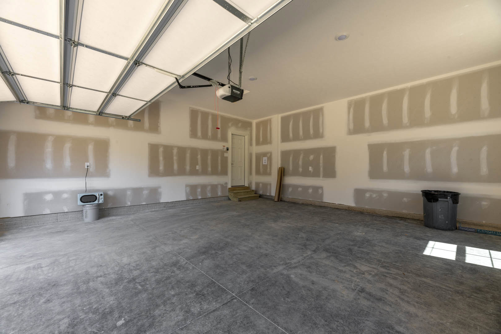 Concrete floor with white ceiling, garage door, interior door, black garbage can, microwave on top of a bucket, and daylight from window