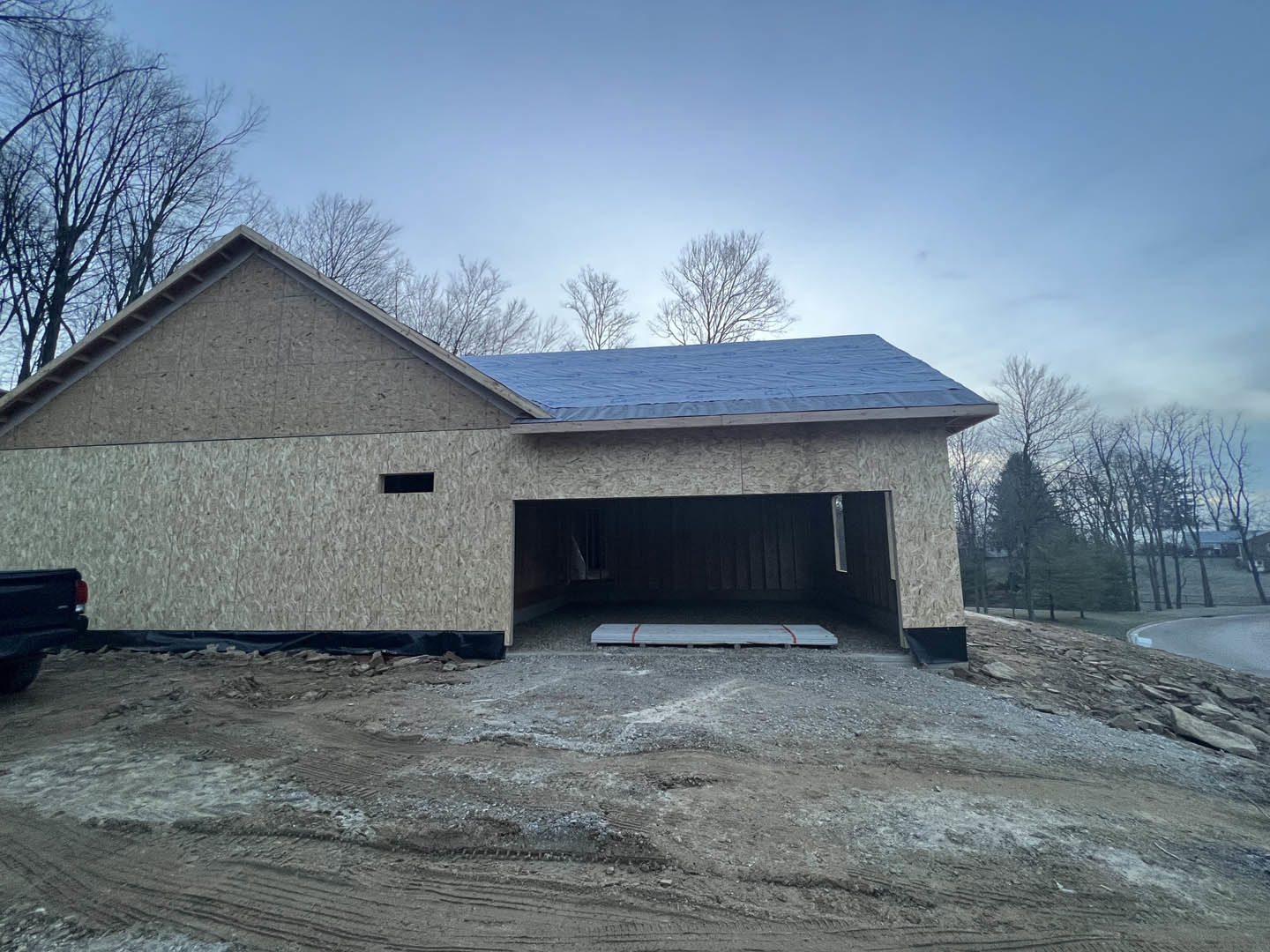 Framed custom home under construction with attached garage, exposed plywood sheathing, dirt ground with tire tracks, leafless tree branches, blue winter sky