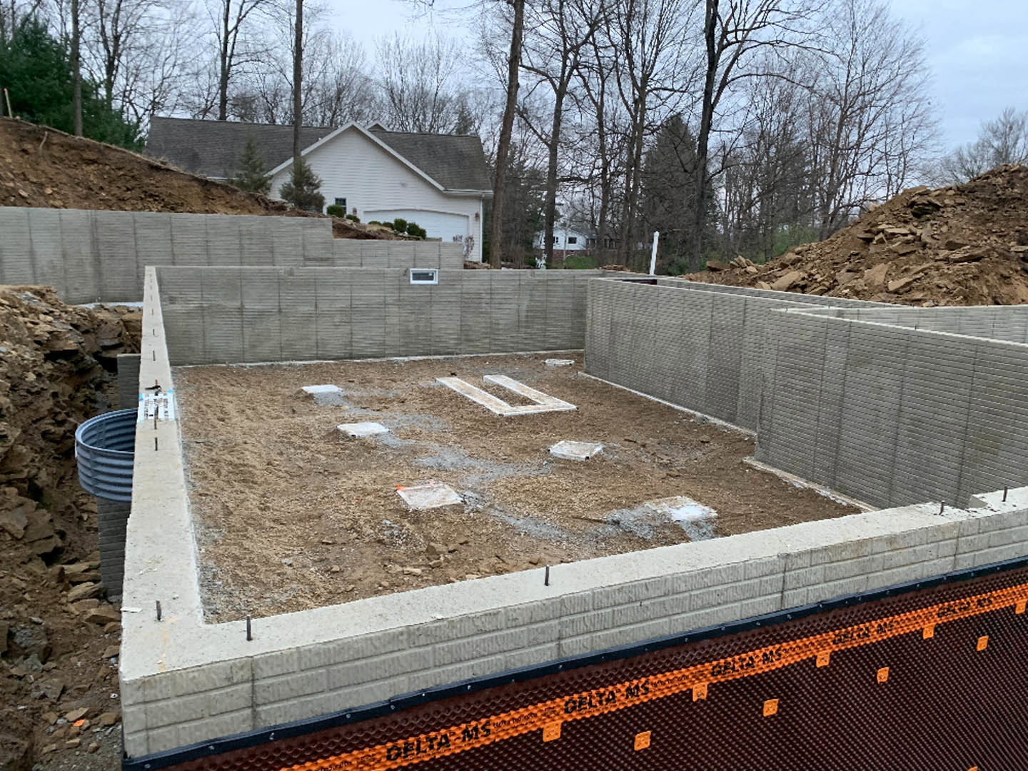 Concrete foundation under construction with exposed rebar, temporary fencing, and adjacent stone wall; white rectangular object and pipe visible on gravel ground, surrounded by