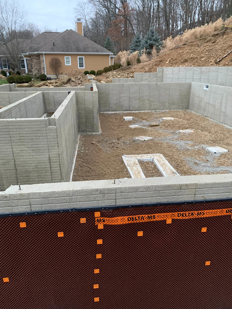 Partially built house with composite siding, visible chimney, surrounded by trees and construction materials on bare ground under a clear sky