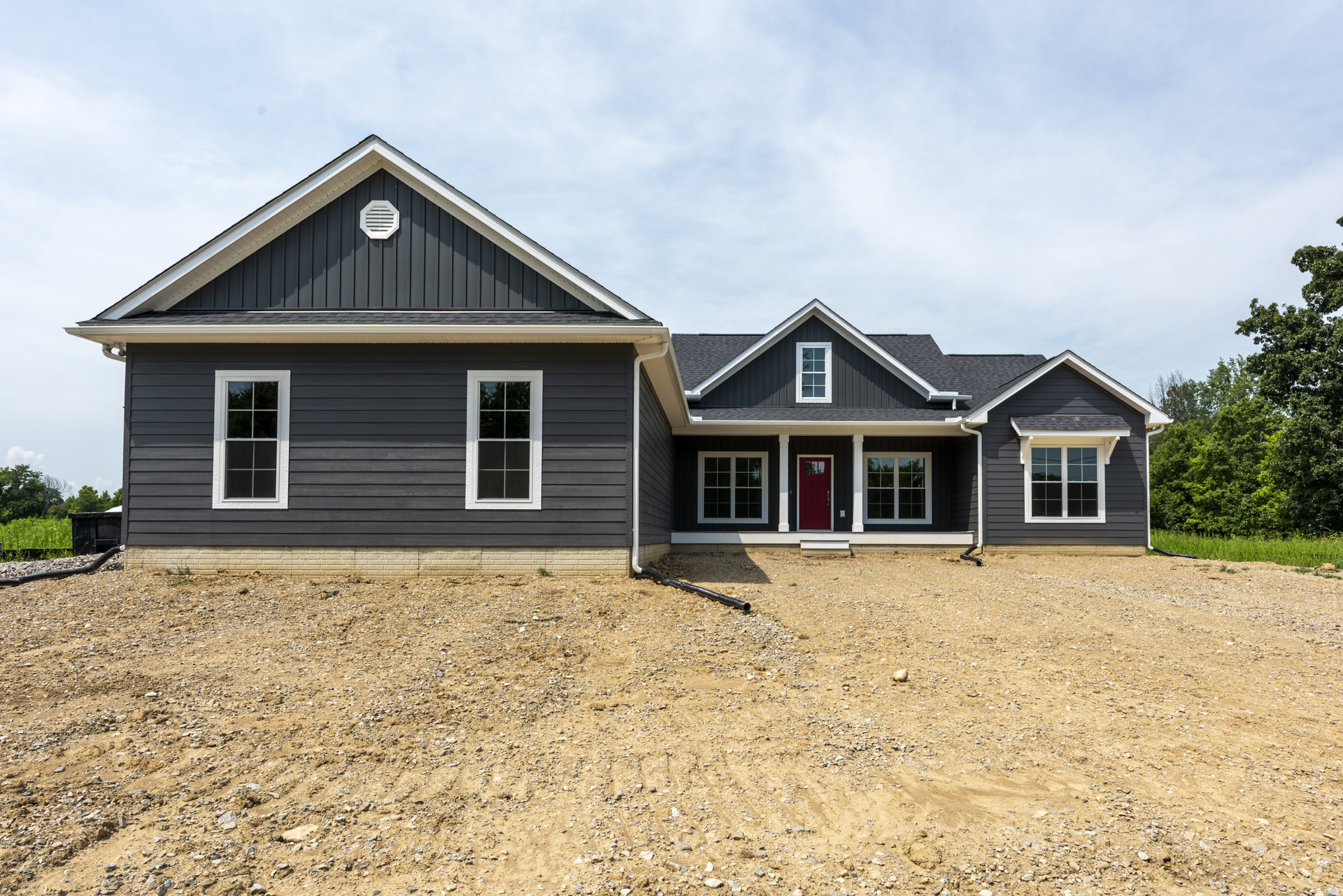 Black exterior wall with white vent, white-framed window, red door featuring glass panel, gravel hill in front, exposed pipe on ground, cloudy sky overhead