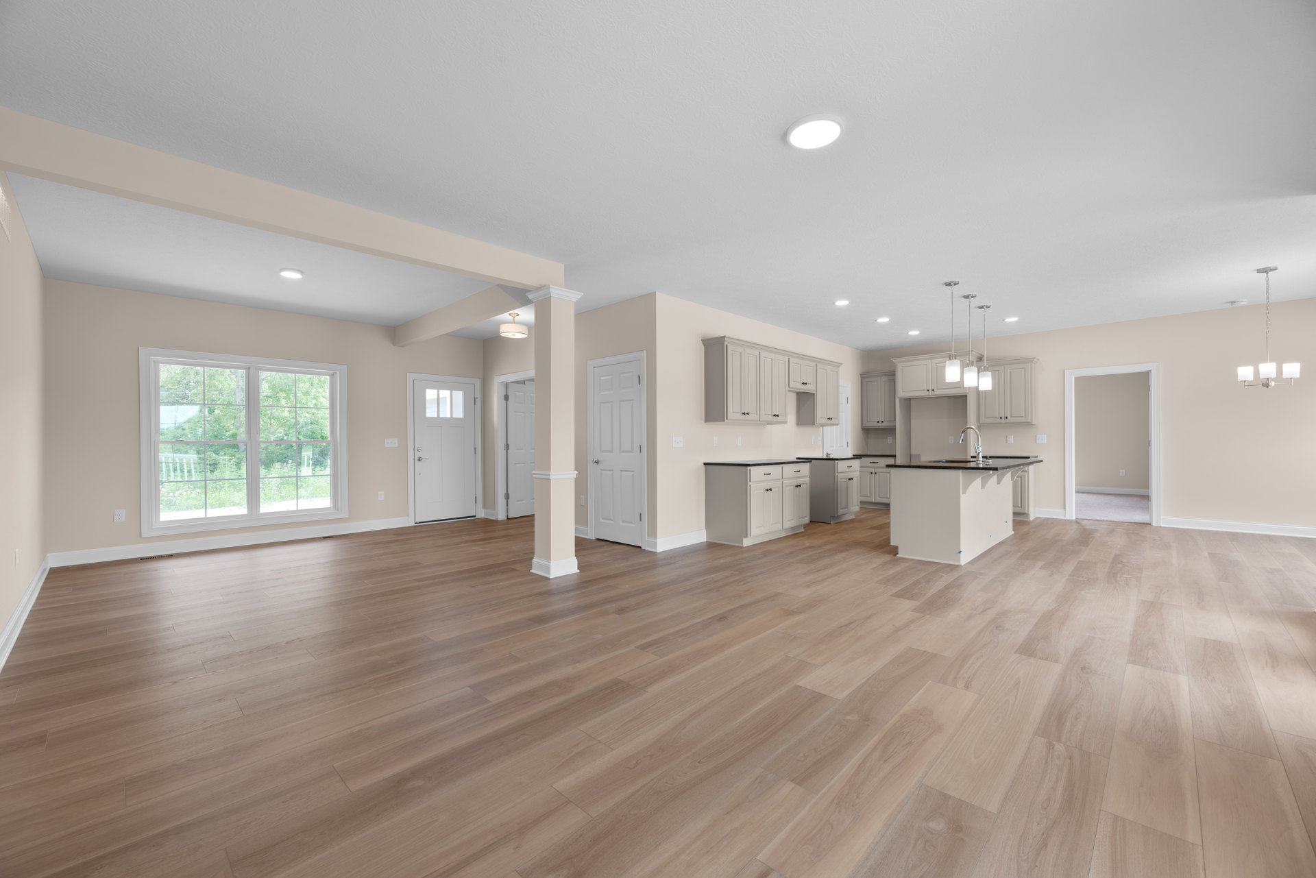 Spacious open floor plan featuring wood flooring, white walls, and white kitchen cabinets; dining area adjacent to a window with trees visible outside, ceiling light fixture, and a