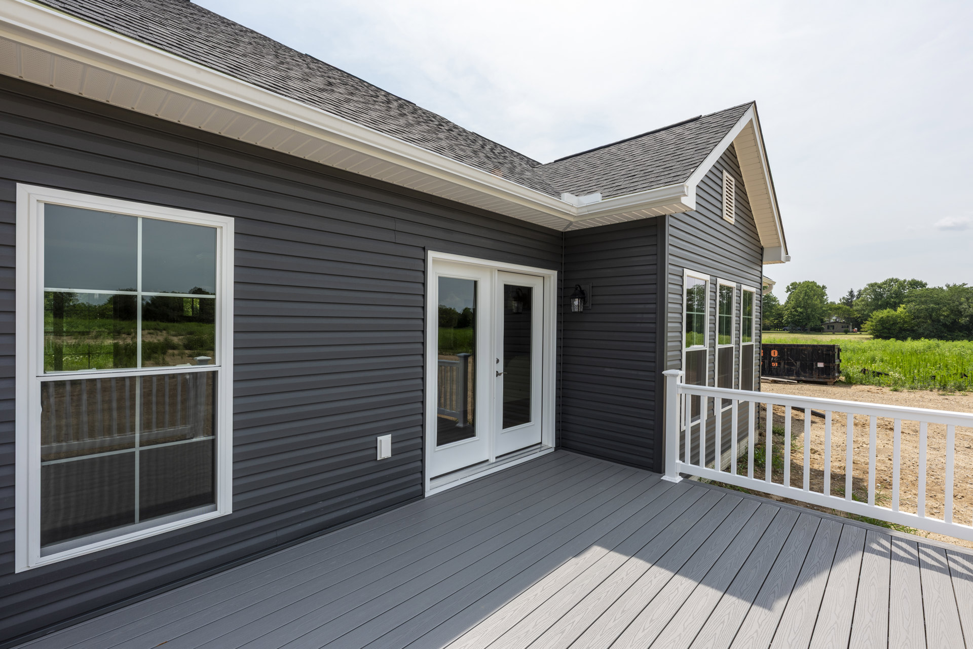 Deck with white railing, white door with glass panels, windows with screens, light-colored siding, black container with orange stickers, outdoor residential setting