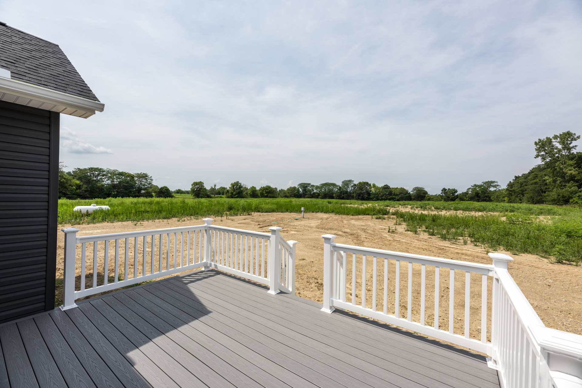 Wood deck with white railings overlooking a cornfield, trees, and a white tank in the distance under a partly cloudy sky