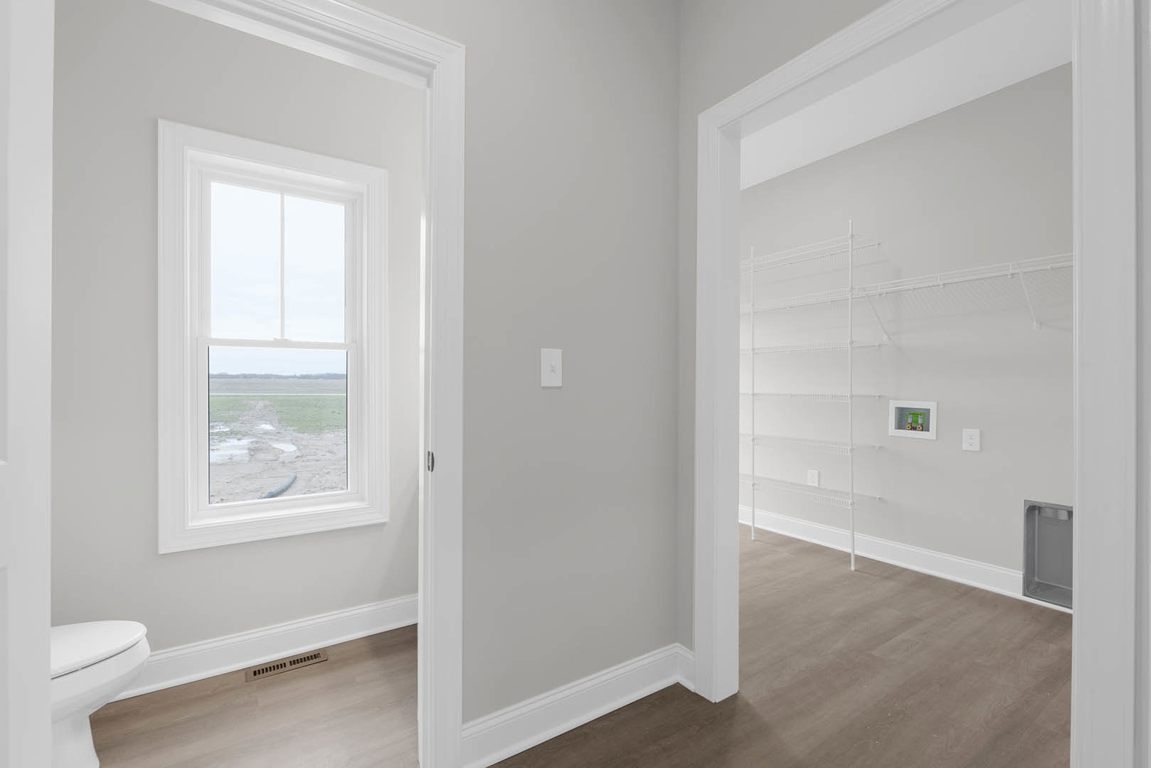 Bathroom with white tile flooring, built-in shelves beside a window overlooking a grassy field, white toilet, vent on ceiling, and white walls with black trim.