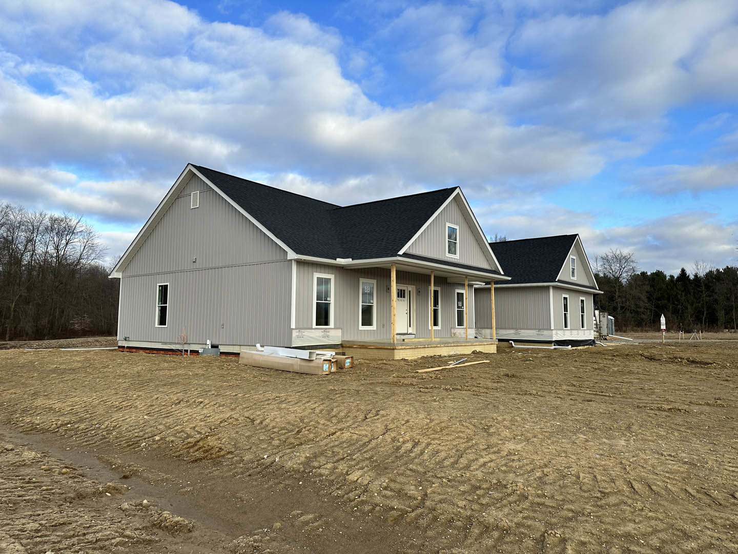 Wood-framed house under construction with exposed metal beam, unfinished porch, surrounded by dirt lot and leafless trees under blue sky with scattered clouds