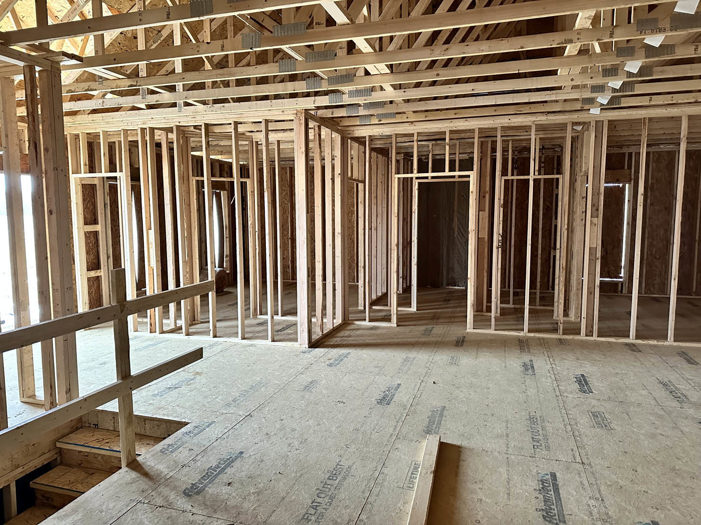 Exposed wood framing and beams inside a house under construction, unfinished floors and staircase, visible lumber and insulation