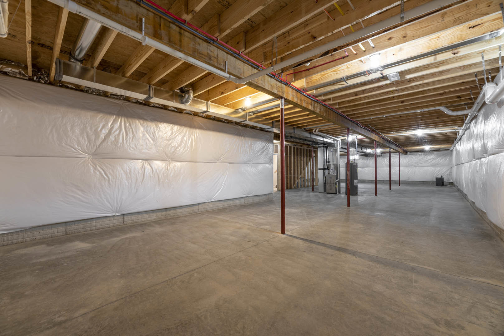Basement room with large white plastic-covered wall, exposed concrete floor, red support pole, ceiling pipes, single light bulb, and close-up of white electrical box.