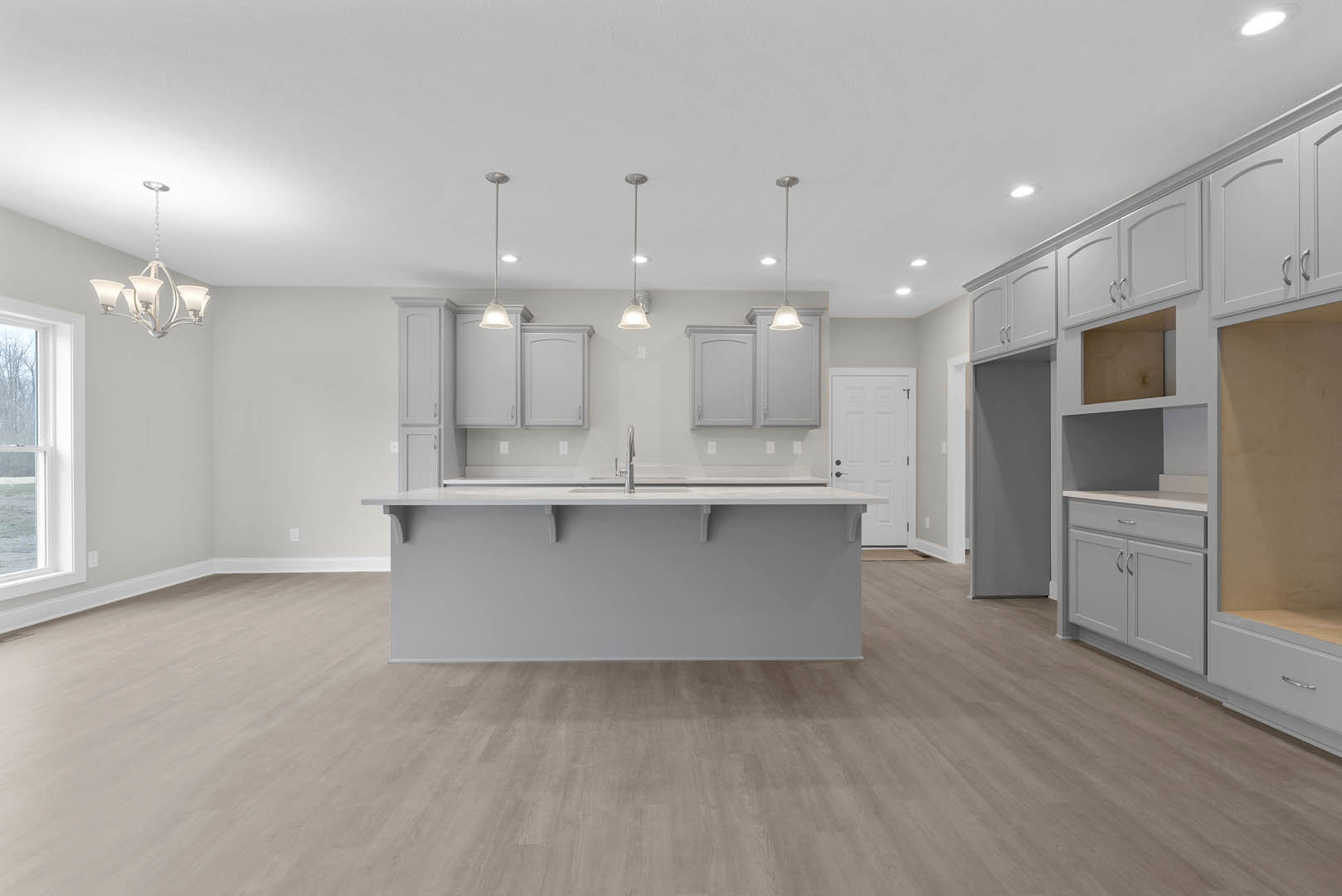 Spacious kitchen featuring a large white island with built-in sink and faucet, wood flooring, white cabinetry with black hardware, white walls, and a window letting in natural