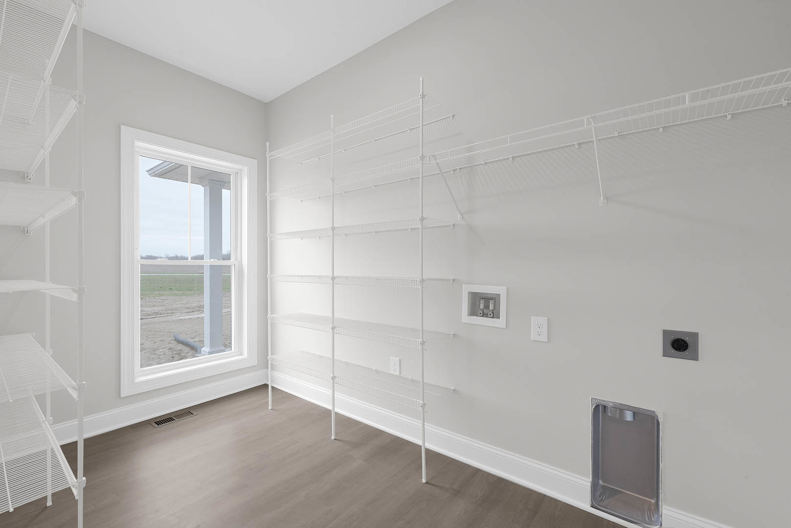 Built-in white shelves along a plaster wall, window overlooking grassy field, light wood flooring, black round outlet on shelf, two small decorative objects in white frame