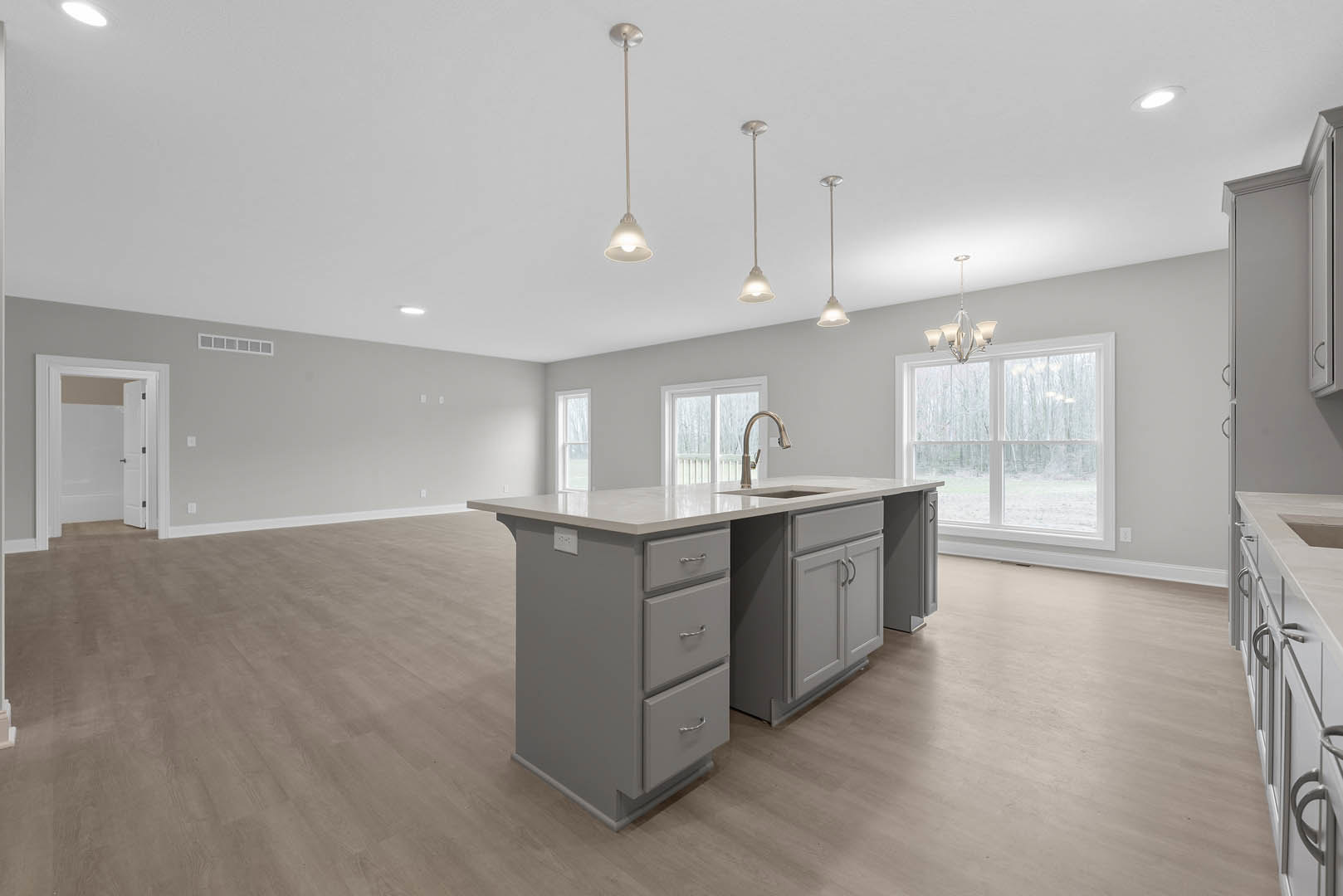 Spacious kitchen featuring a large central island with drawers, light wood flooring, white cabinetry, stainless steel sink and faucet, window with pendant light fixture, white door