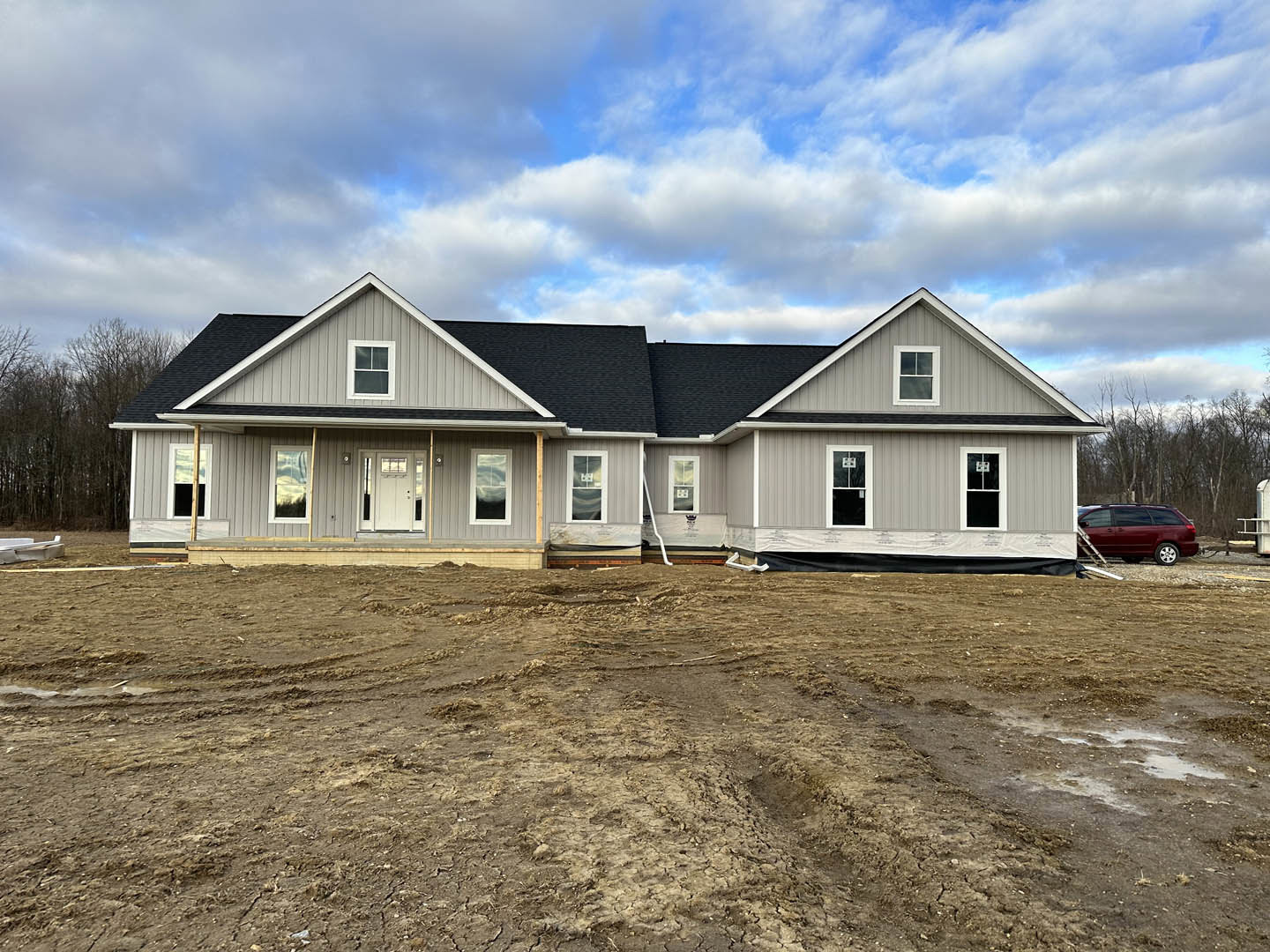 Partially built house with exposed framing, white window frames, dirt construction site, scattered white pipes, red van with ladder, cloudy blue sky overhead