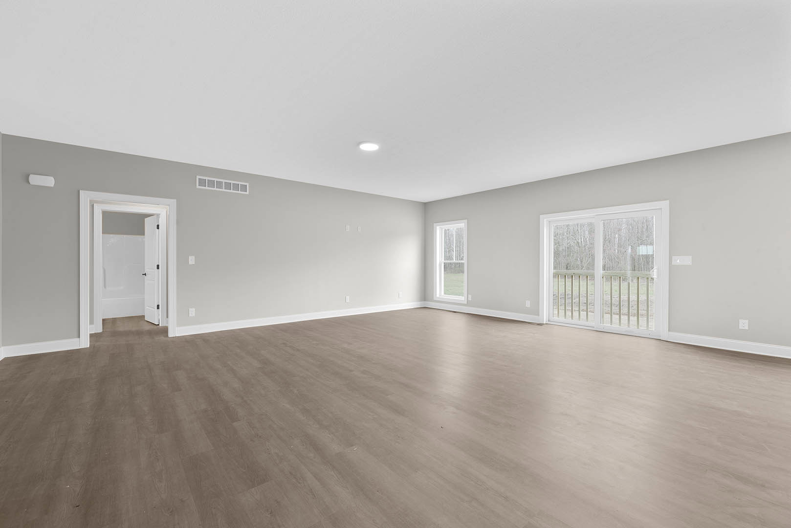Spacious room with light wood flooring, white walls, sliding glass door leading to a balcony with railing, large window offering outdoor view, and white door with black hardware