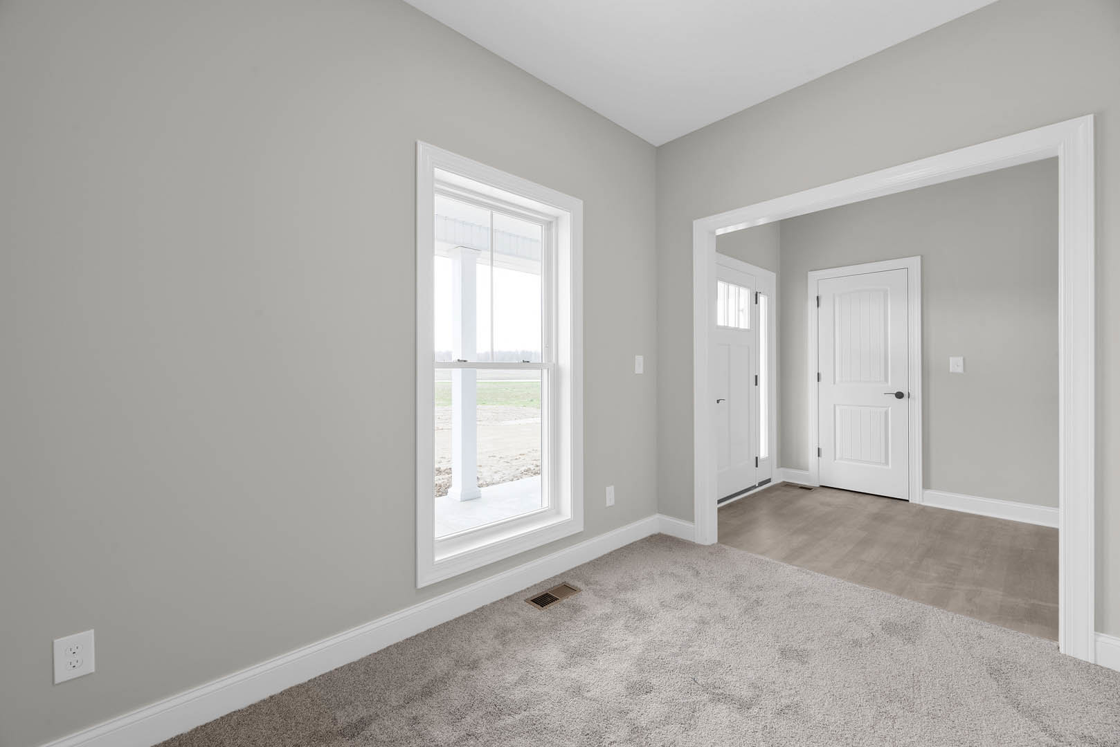 Carpeted room with white walls, large window, white door with black handle, wall vent, and electrical outlet