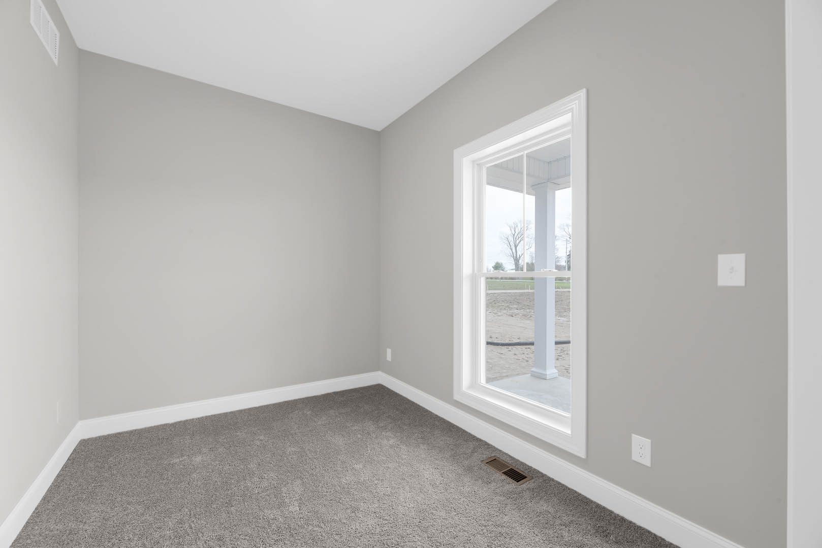 Neutral-toned carpeted room with white walls, large window overlooking grassy field, metal floor vent, and wall outlet visible near baseboard molding
