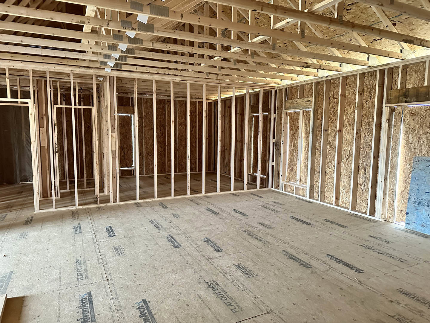 Unfinished room with exposed wood beams, plywood subfloor marked with black text, partially installed ceiling insulation, and visible wood framing