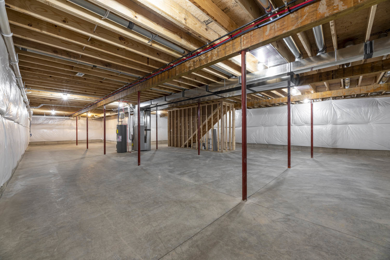 Basement room with exposed steel beam and metal box, concrete floor with red support poles, pipes along unfinished ceiling, white sheet covering part of wall, wooden staircase