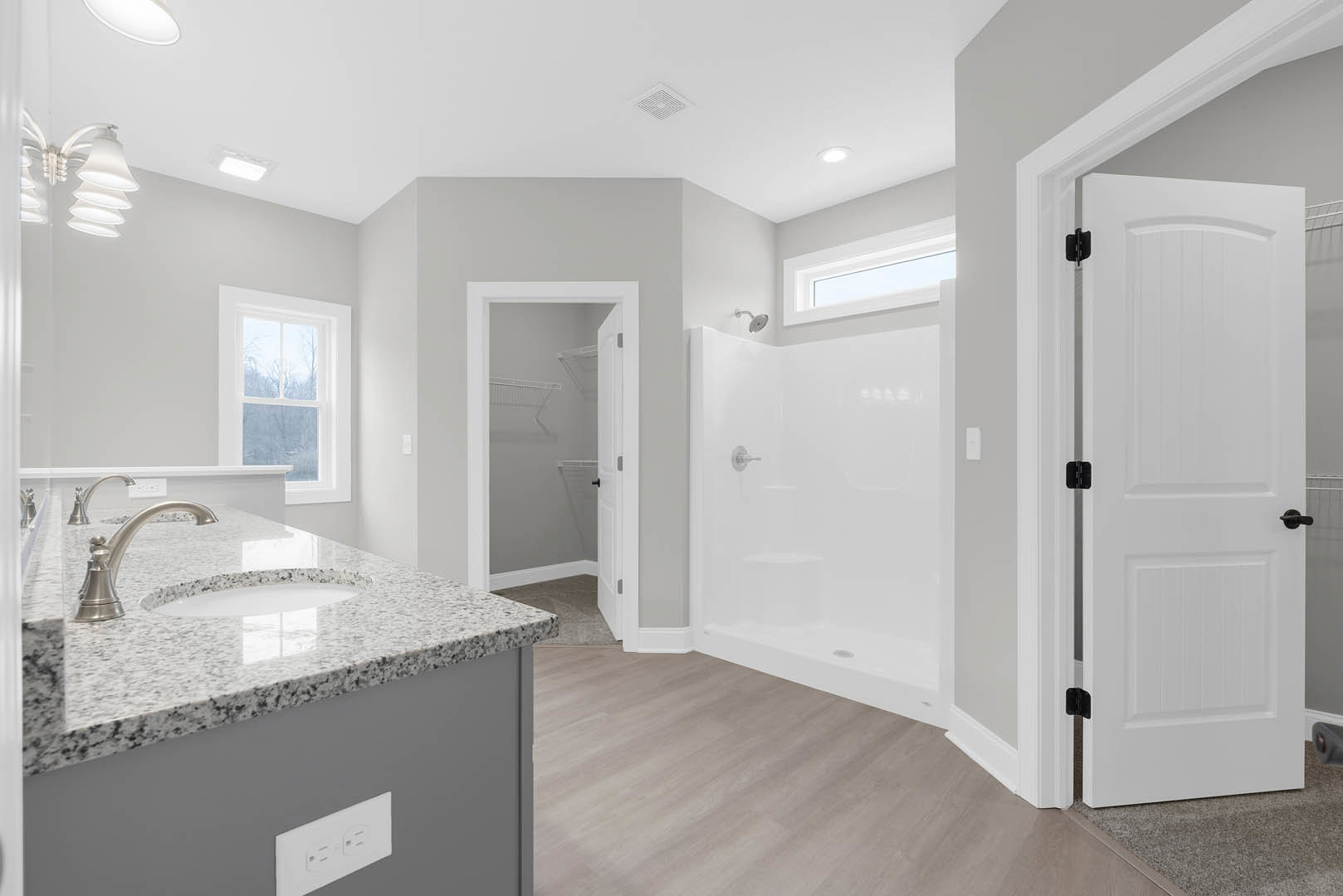 Bathroom with white tile floor, built-in shower, white sink and countertop, black hardware on white door, window overlooking trees, open closet door, cabinetry with modern plumbing