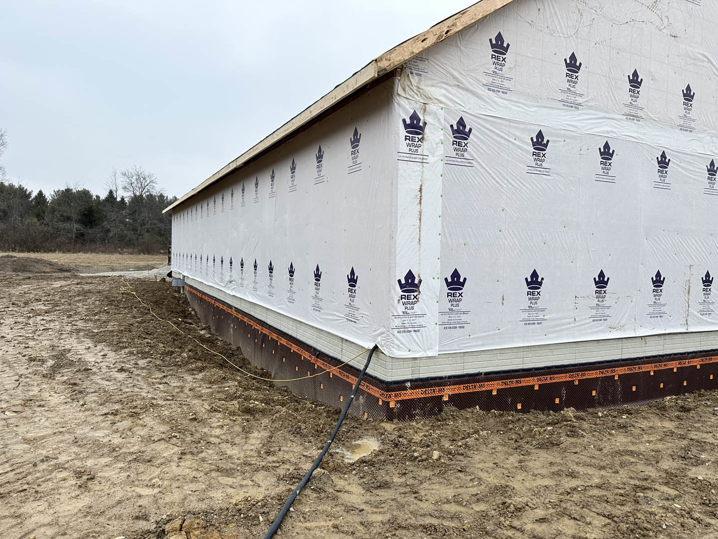 White plastic sheeting covers exterior walls of a custom home under construction, with black hose and pipe visible near foundation, surrounded by trees and dirt ground.