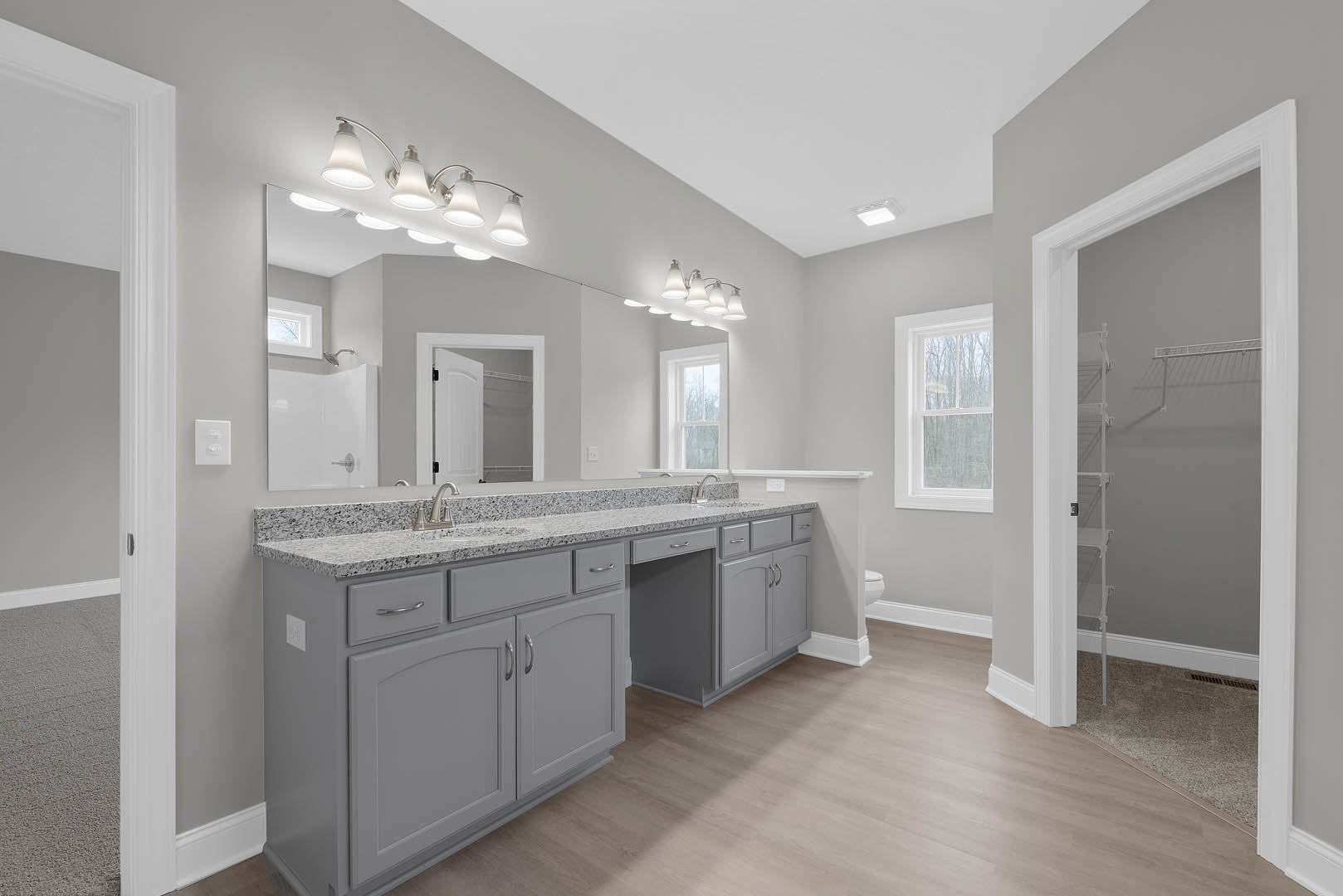 Bathroom with marble countertop, under-mount sink, tile floor, white cabinetry, multi-bulb light fixture above mirror, window showing trees, open white door leading to closet