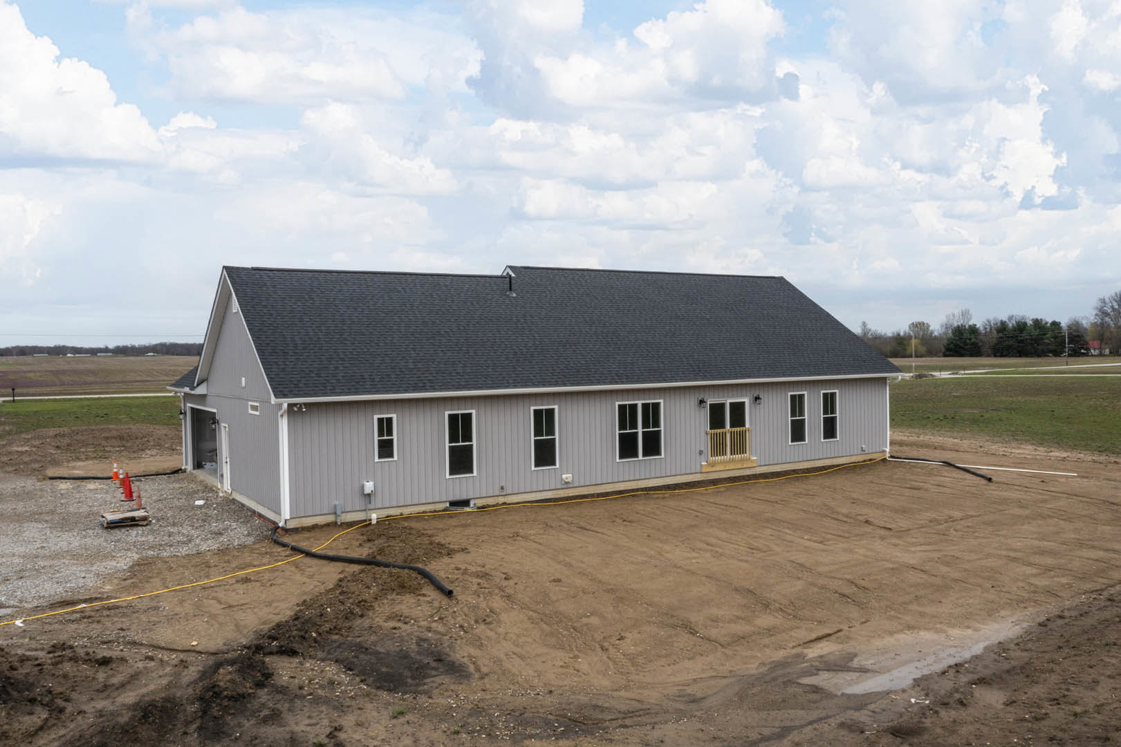 Grey house under construction with black roof, white-framed window, exposed dirt lot, and cloudy sky