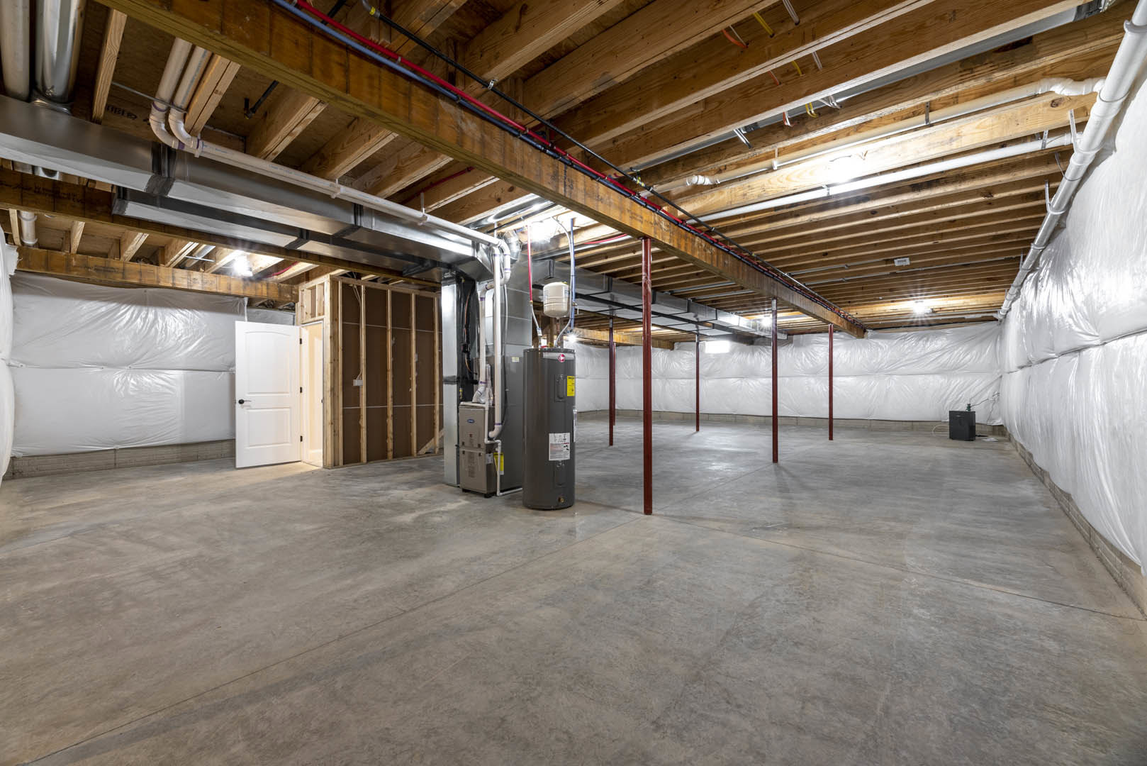 Basement room with exposed steel beam, unfinished wood framing, wooden ceiling with visible pipes, large grey cylinder, and white door with black handle