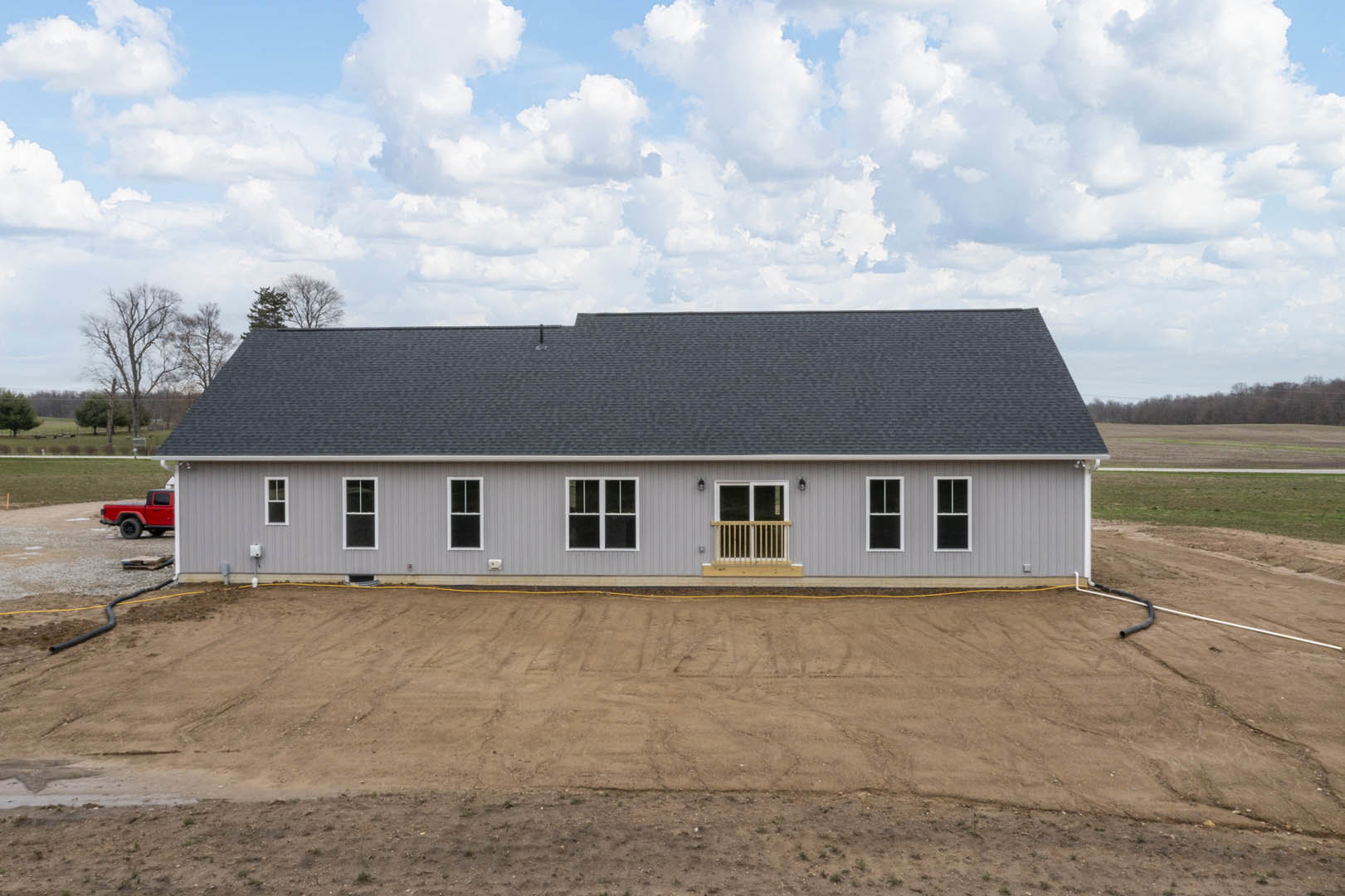 Grey two-story house with balcony, dark-framed windows, and pitched roof under cloudy sky; red truck parked on rocky dirt area with yellow hose; sparse landscaping and trees in