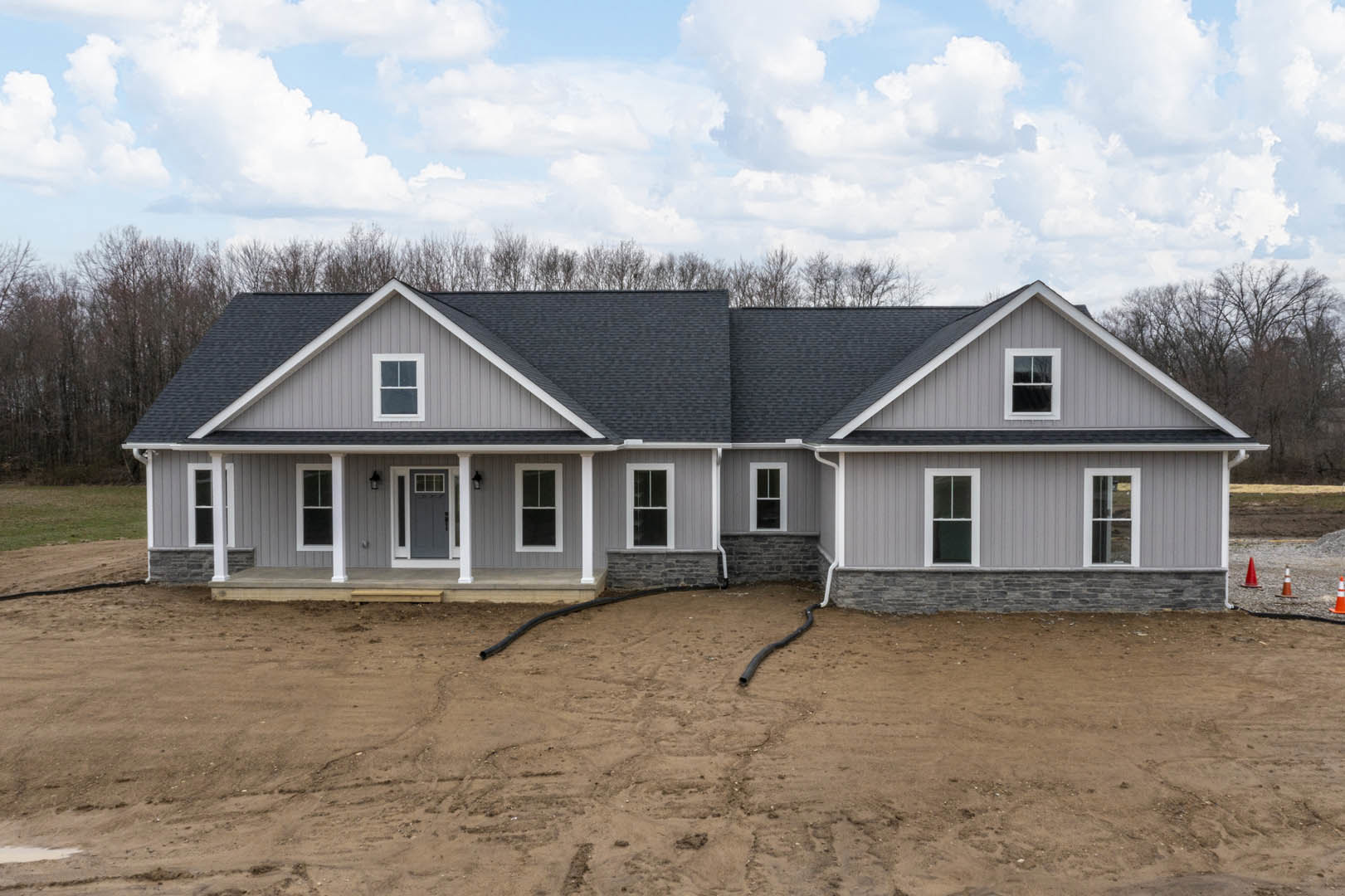 Partially built house with grey roof, white-framed windows, exposed dirt yard, garden hose in front, surrounded by trees under cloudy sky