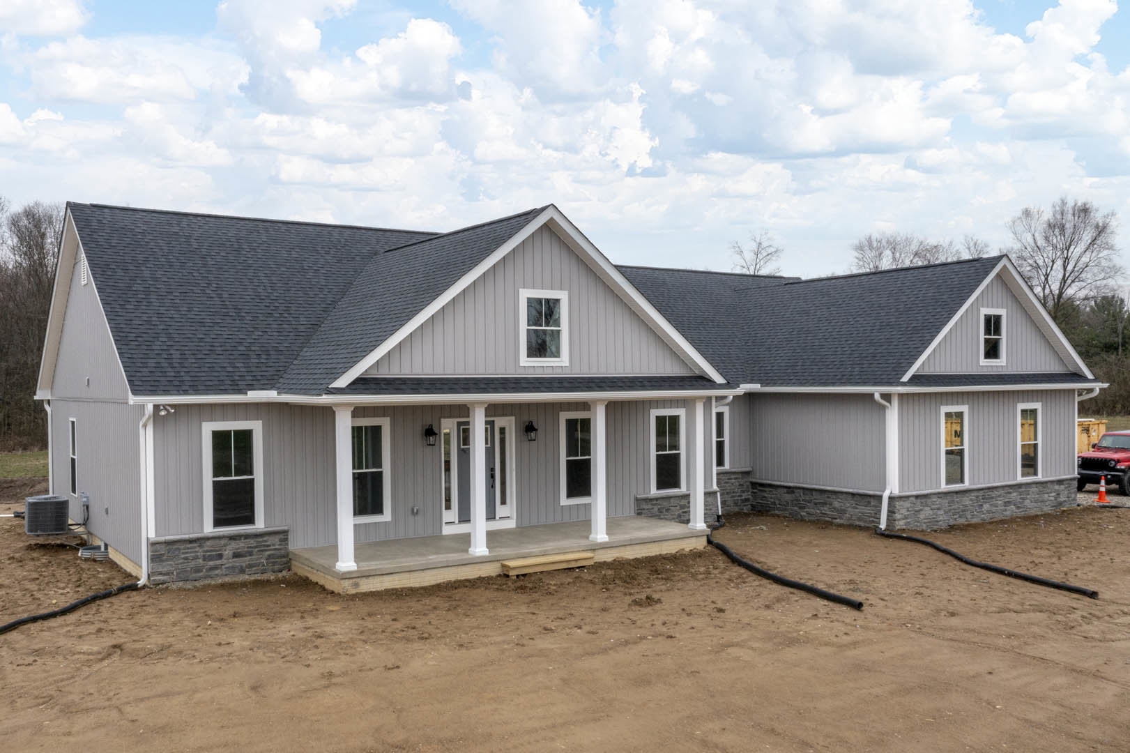 Two-story home with white-framed windows, covered porch, dirt yard, red car parked roadside, and large metal utility box on ground
