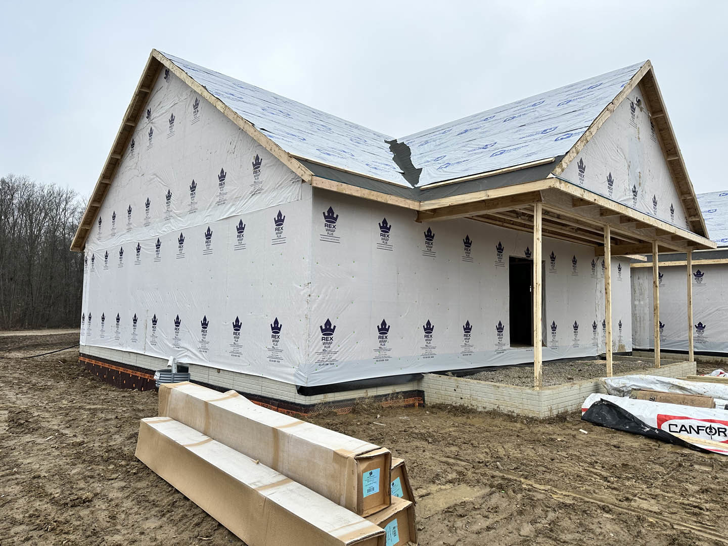 Partially built house with exposed wooden framing, scattered lumber on dirt ground, unfinished roof, and surrounding trees under a clear sky