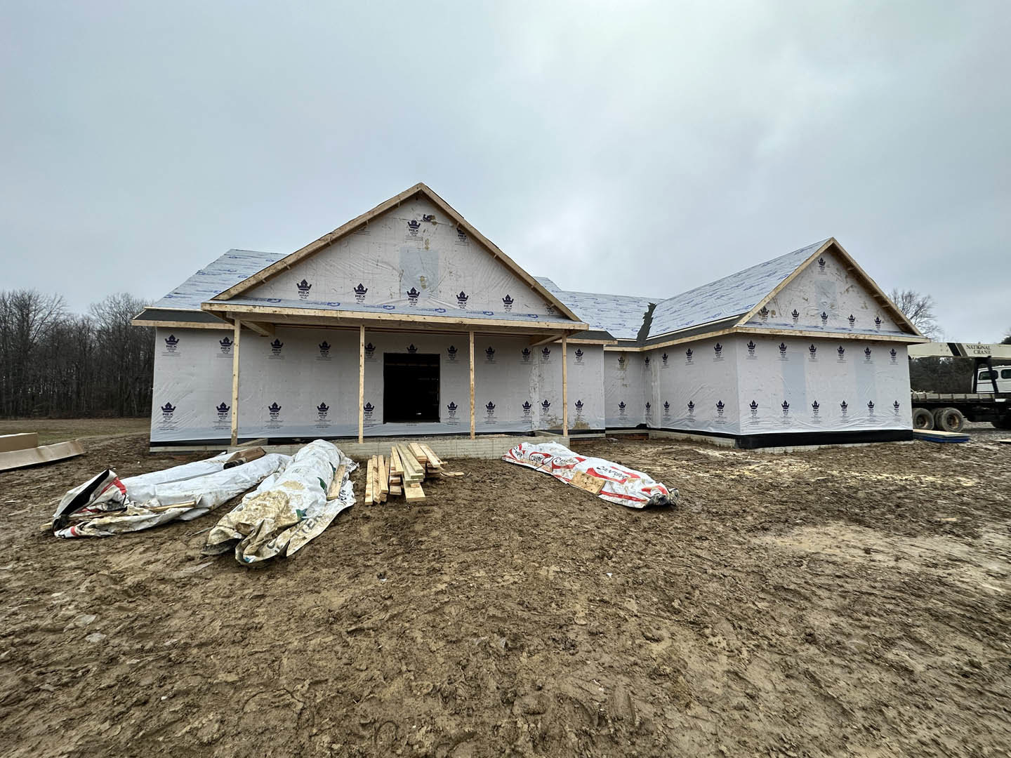 Partially built house with exposed roof trusses, black front door, exterior light fixture, pile of sand, stacked wood planks, white and red construction bag, dirt yard, cloudy sky