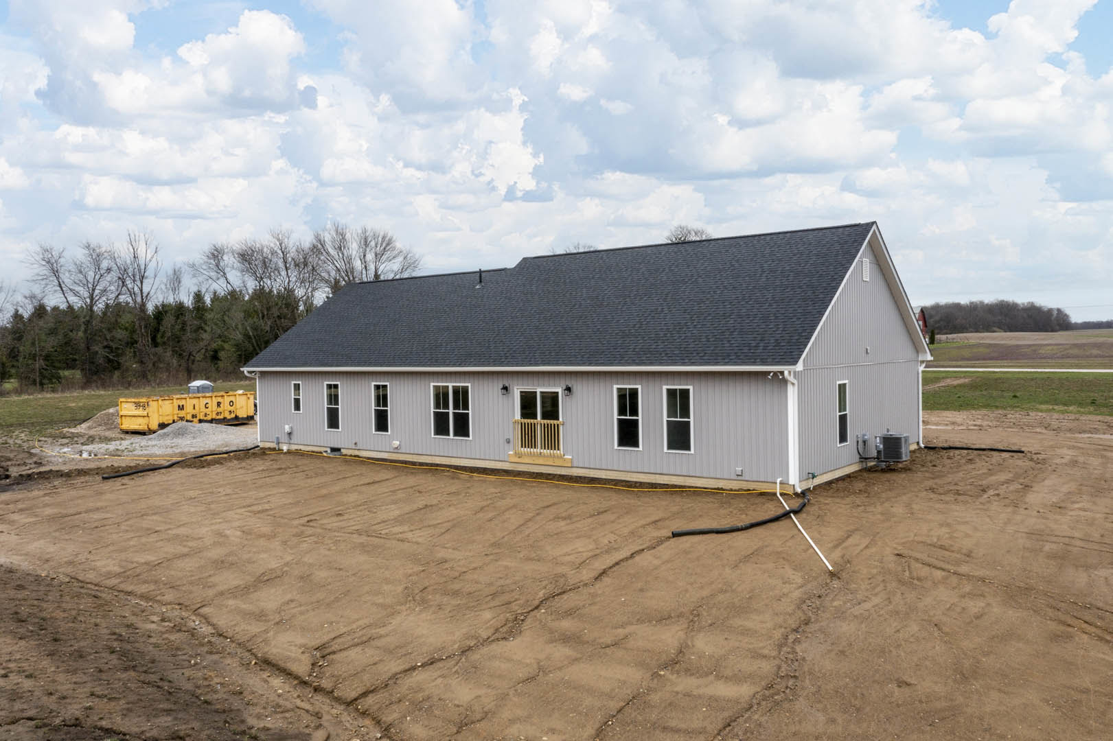 Grey two-story house with balcony, metal container labeled in black, unfinished dirt yard, trees and clouds in background