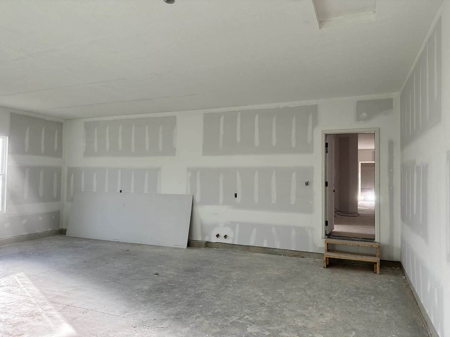 White plaster wall and ceiling, concrete floor, wooden bench near door with sunlight streaming in, person standing beside wall with visible paint drips