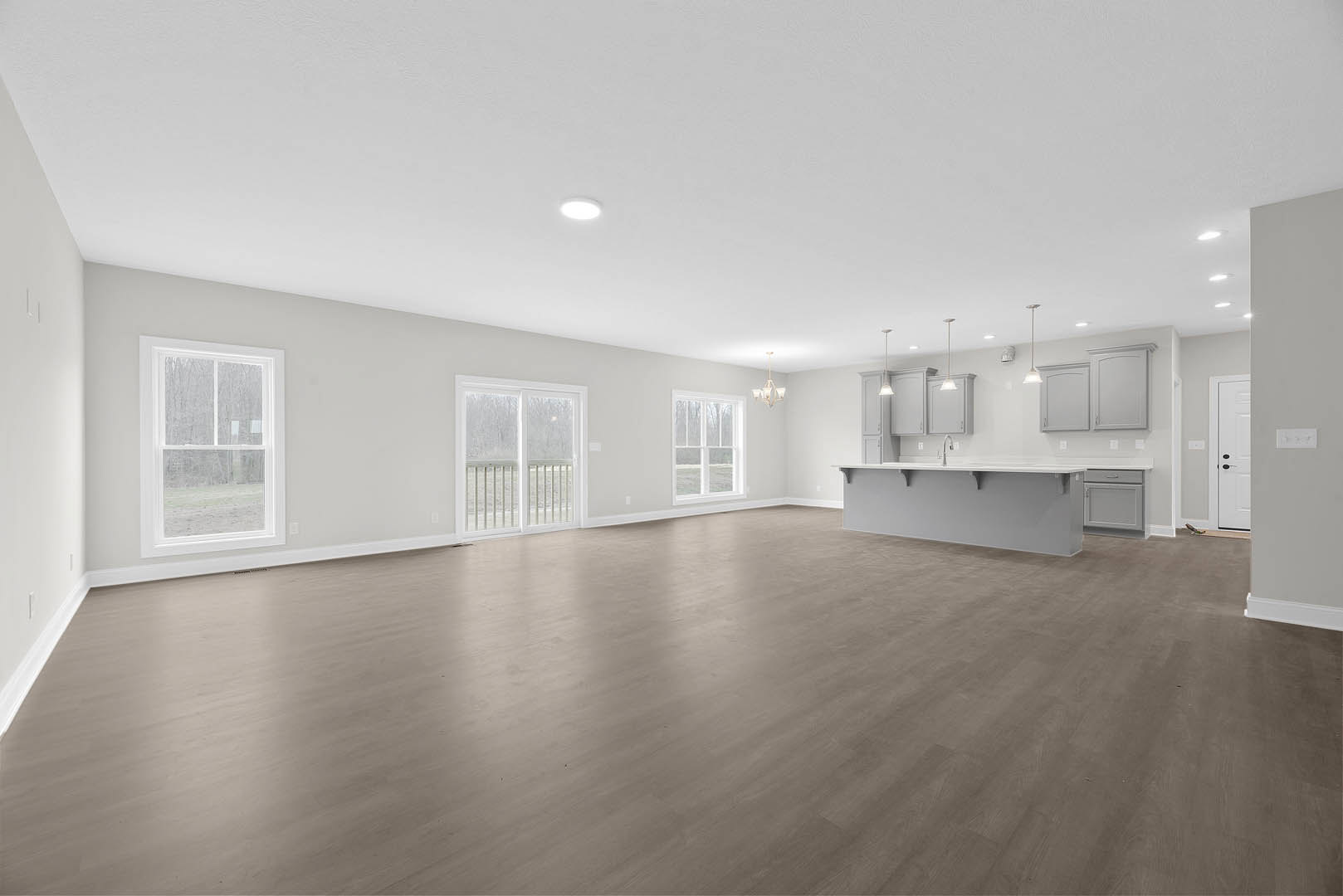 Spacious open-plan room featuring wood flooring, modern white kitchen cabinets, white shelving, and large window with white frame overlooking exterior view.