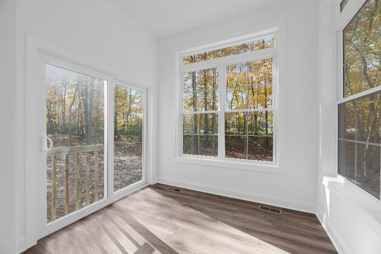 Open living area with wide plank wood flooring, large windows overlooking leafy trees, sliding glass door leading to deck, and vent in floor corner.