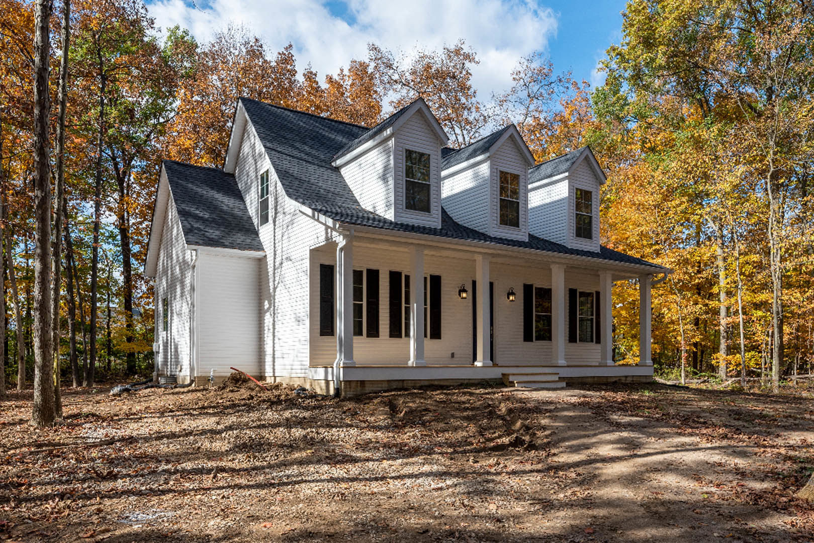 White siding house with black shingle roof, large windows, dirt yard scattered with leaves, surrounded by mature trees under partly cloudy sky