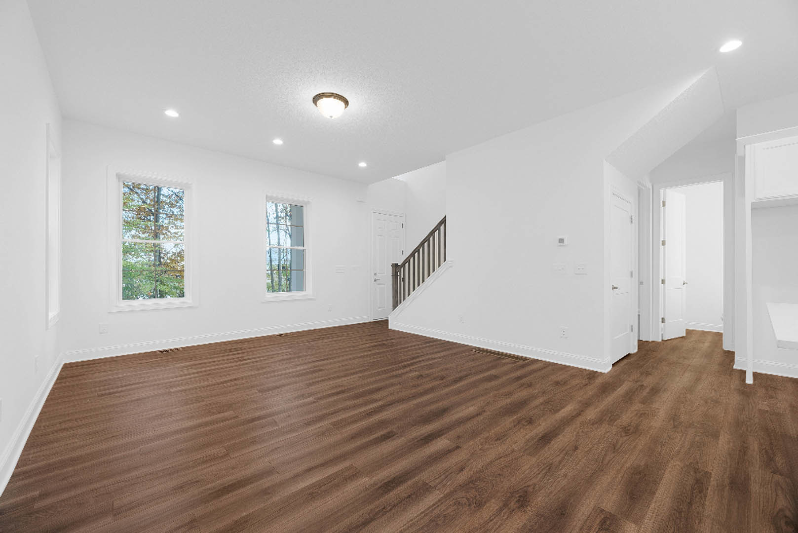 Open living area with wide-plank wood flooring, white plaster walls, modern staircase with wooden treads and metal railing, large window overlooking leafy trees, recessed ceiling