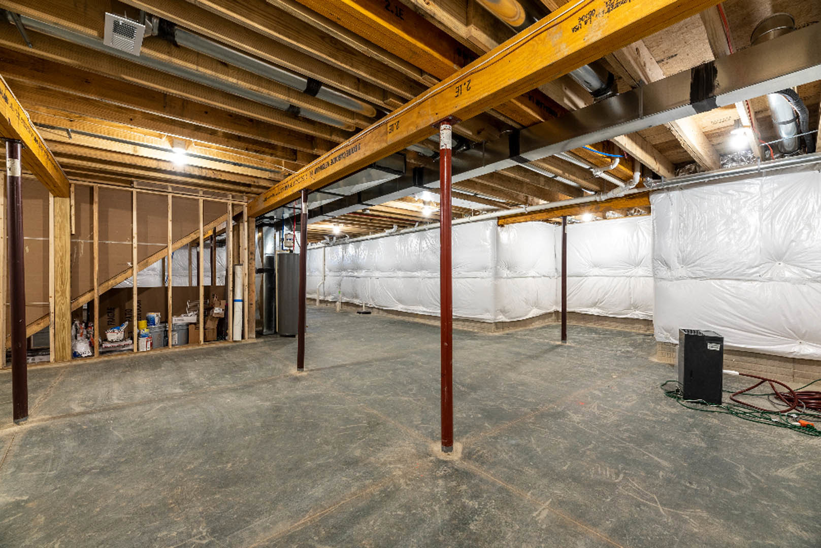 Open room featuring exposed wood beam, white insulated wall, red steel support pole, black electrical box with white label, visible cable, and white square panel with black grid
