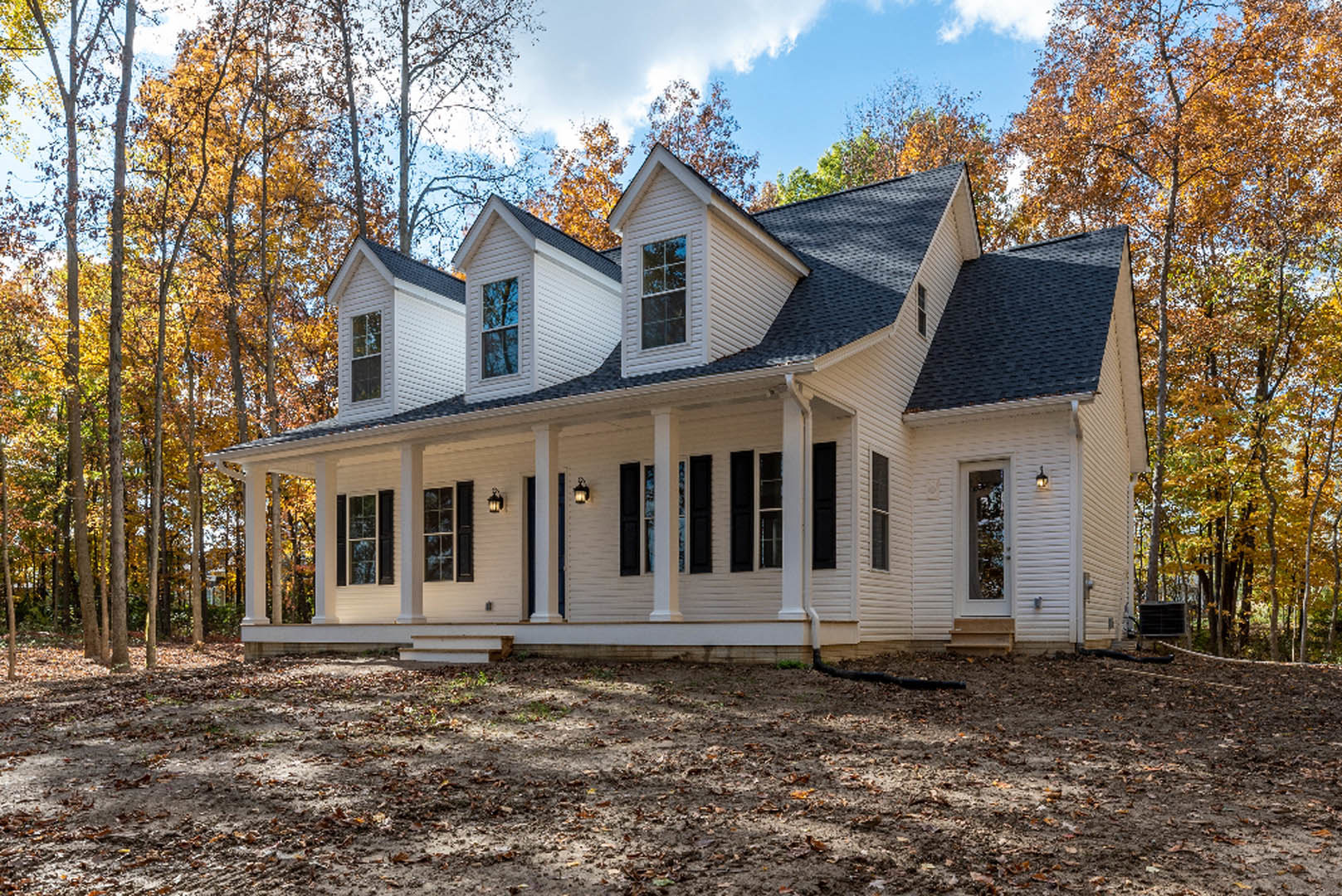 White siding house with black trim and shutters, covered front porch, autumn leaves scattered on dirt yard, large windows with white frames, tree visible outside.