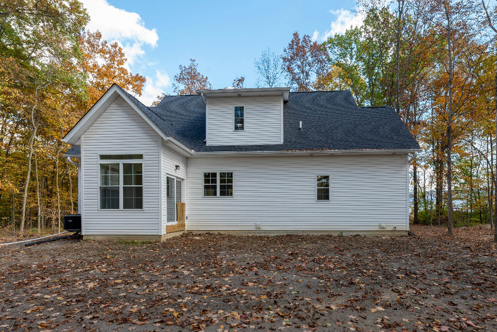 Black-roofed house with white-framed window, gray siding, and autumn leaves scattered on the ground near the exterior wall