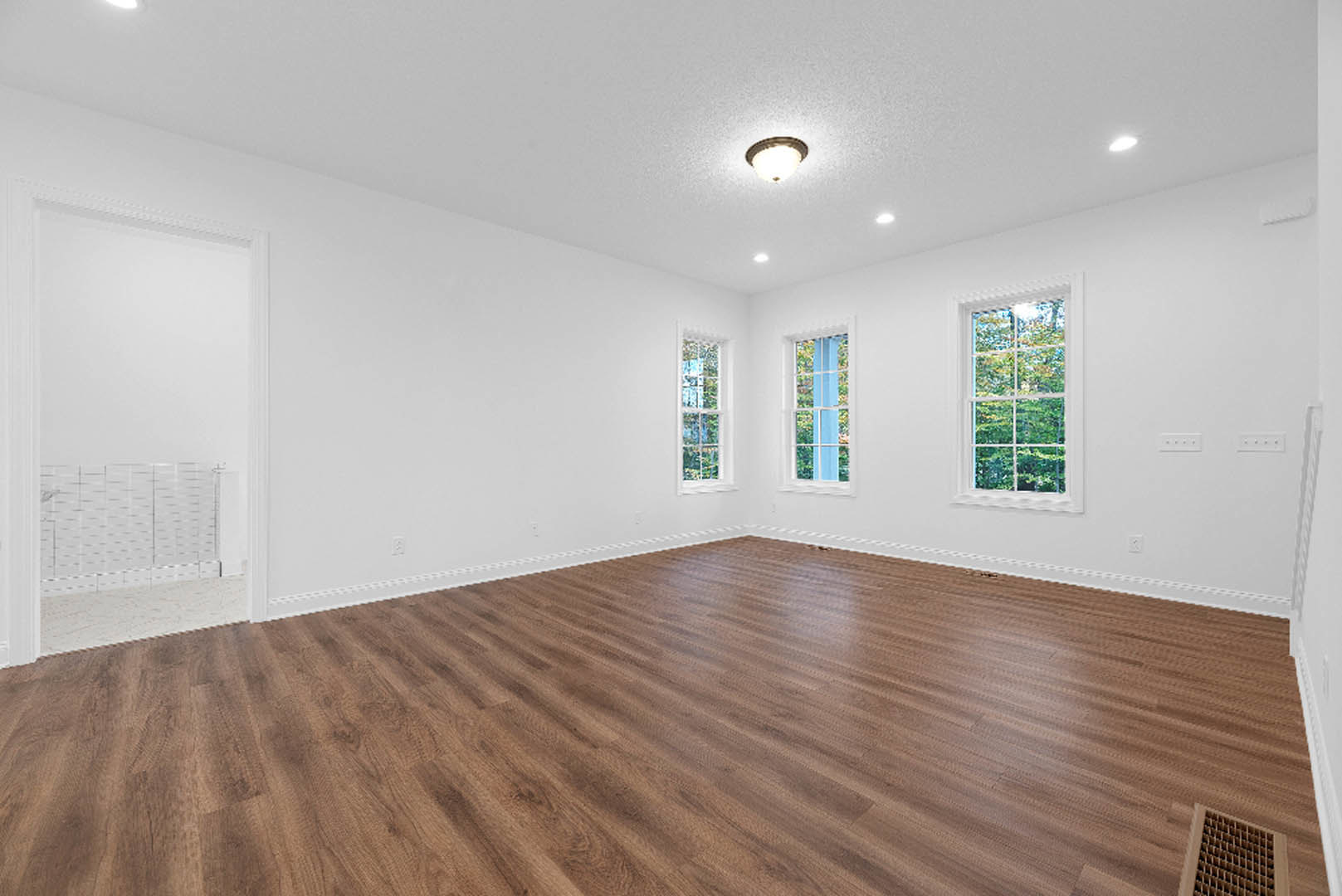 Spacious room with hardwood flooring, large windows overlooking trees, white plaster walls, ceiling light fixture, and a vent near the floor; man standing by the wall.