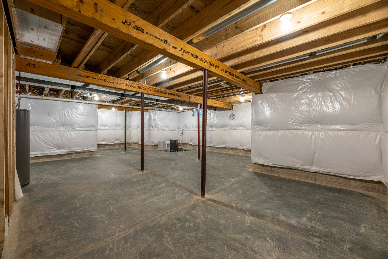 Unfinished basement room with exposed wooden ceiling beams marked with black writing, concrete floor, and walls covered in white plastic sheeting for insulation
