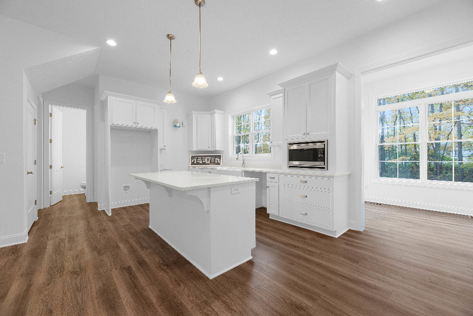 White kitchen with shaker cabinets, wood flooring, white island, stainless microwave, pendant light fixture, window overlooking trees, flagpole visible outside