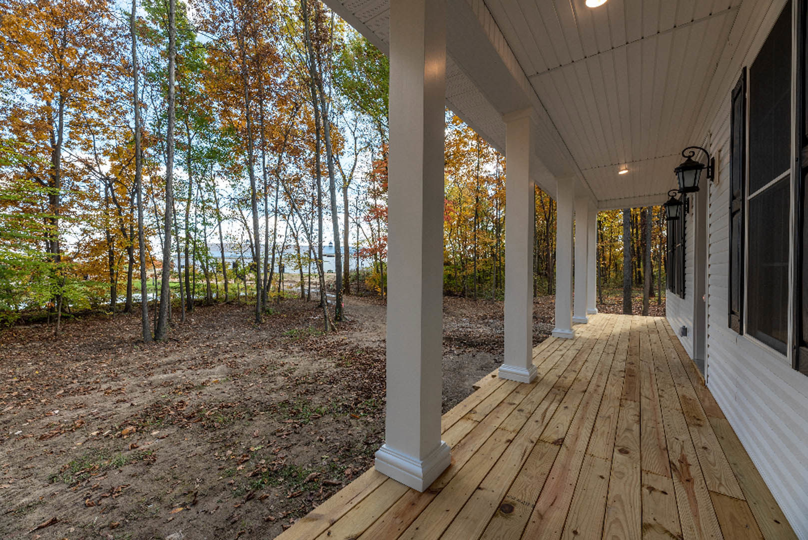 Wooden porch with white pillars and ceiling, bordered by a dirt field scattered with autumn leaves, trees with yellow and green foliage in the background, close-up window visible