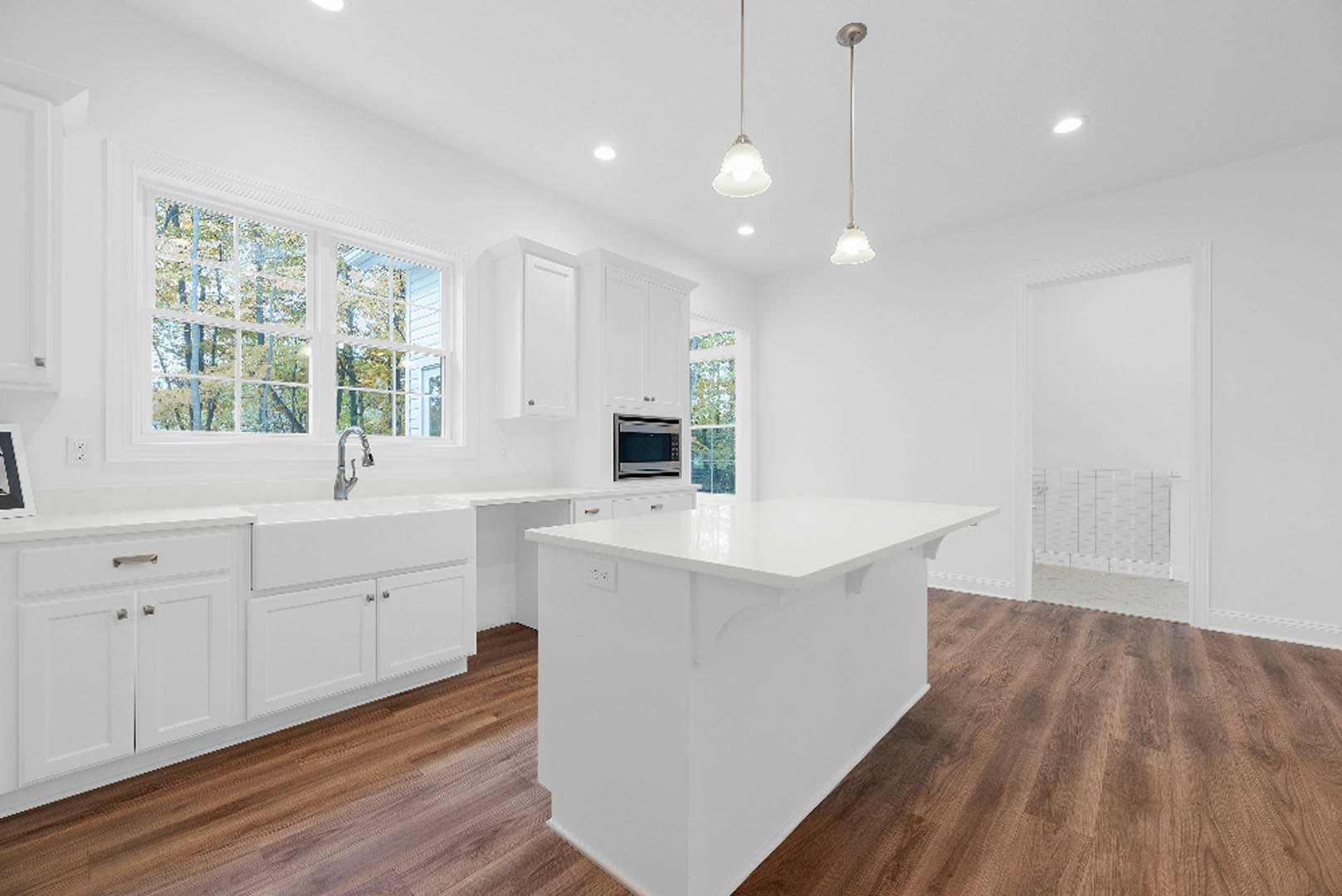 White kitchen with shaker cabinets, wood flooring, central island, stainless microwave, chrome faucet, large window overlooking trees, black and white flag on white pole.