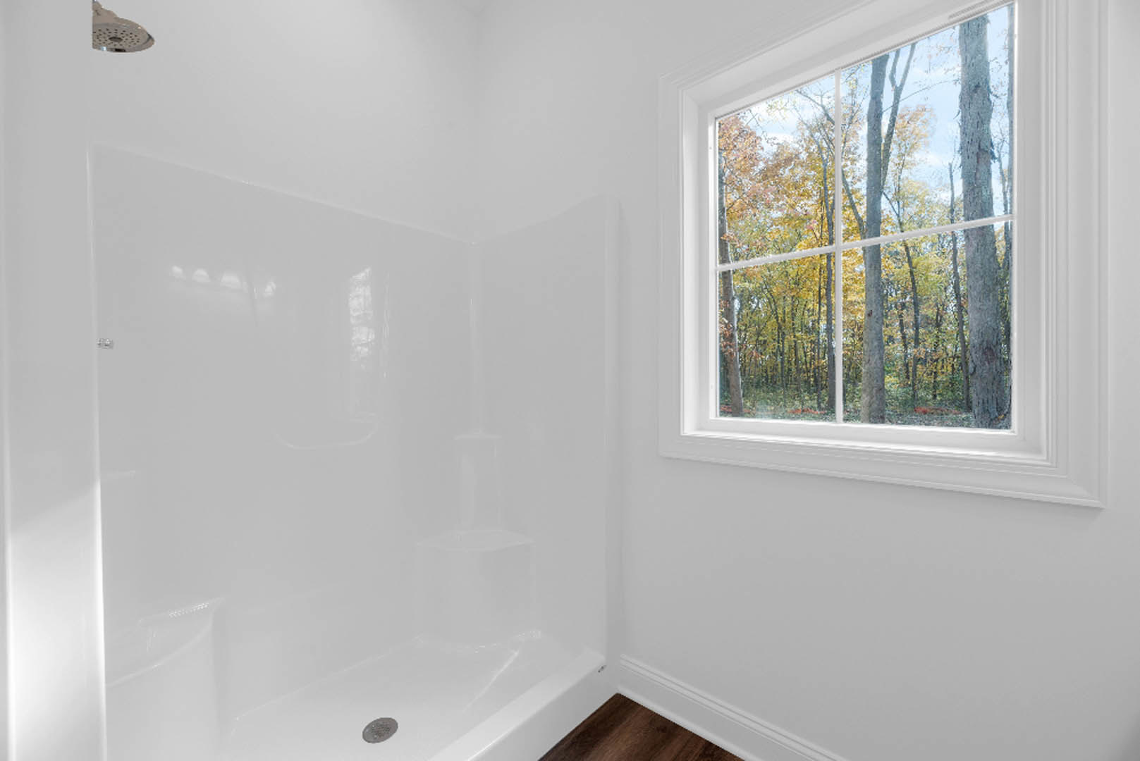 White bathroom with plaster walls, large window overlooking green trees, walk-in shower with round chrome showerhead, light-colored floor.