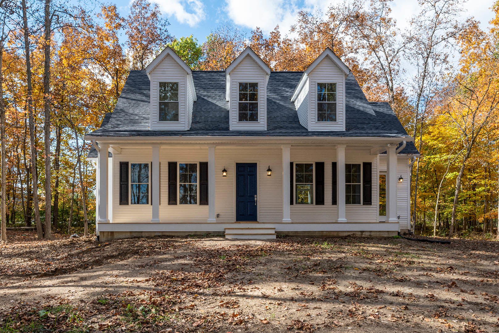 Two-story house with white siding, blue front door, black side door, multi-pane windows reflecting surrounding trees, autumn leaves scattered on ground, mature trees in background