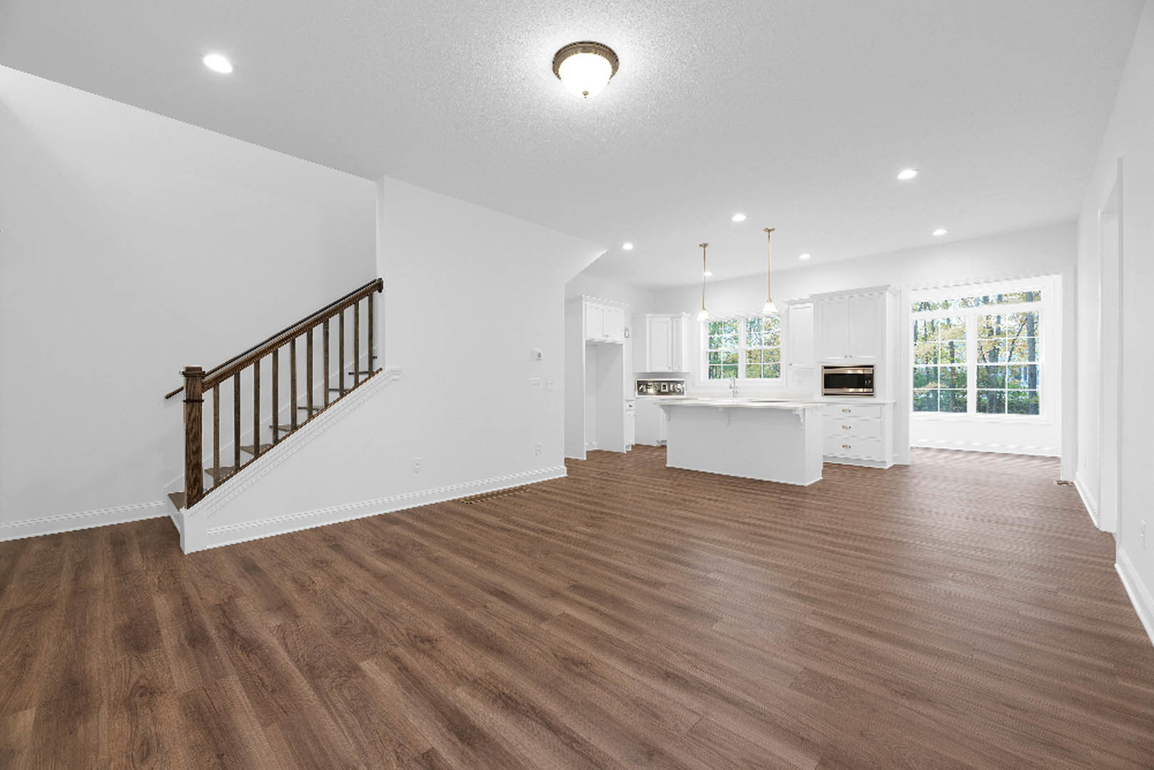 Open-concept room featuring wood flooring, white walls, a staircase with wooden steps, white kitchen counter, ceiling light fixture, and a microwave on a brown surface
